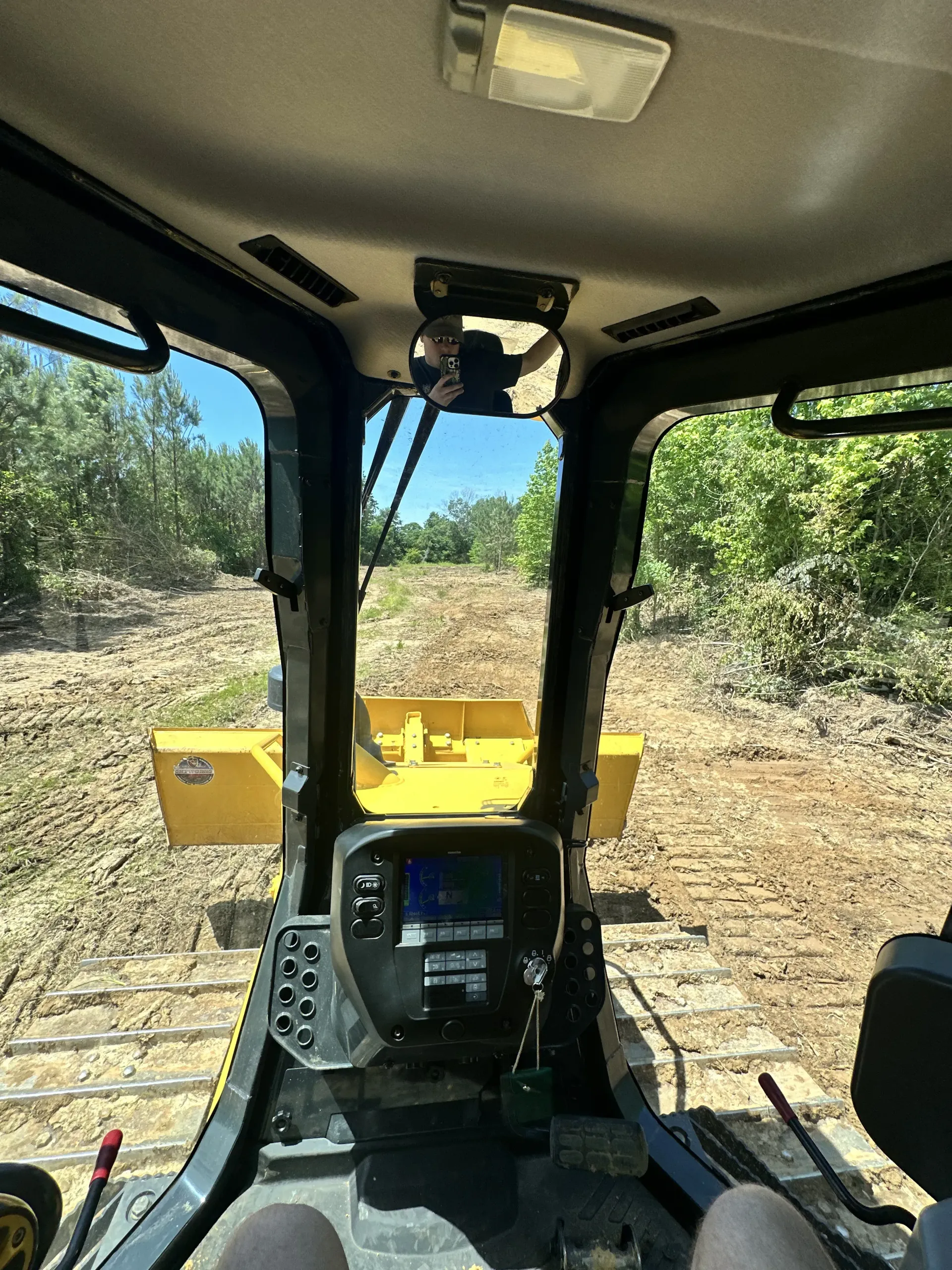View from inside the cab of a yellow bulldozer clearing a wooded area on a sunny day.