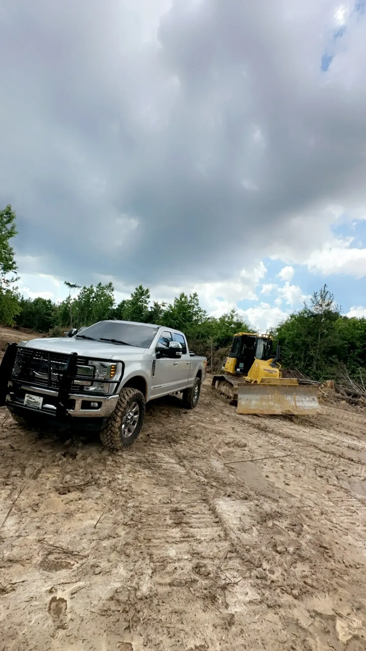 A silver pickup truck with a black brush guard parked on a dirt lot next to a yellow bulldozer under a cloudy sky.