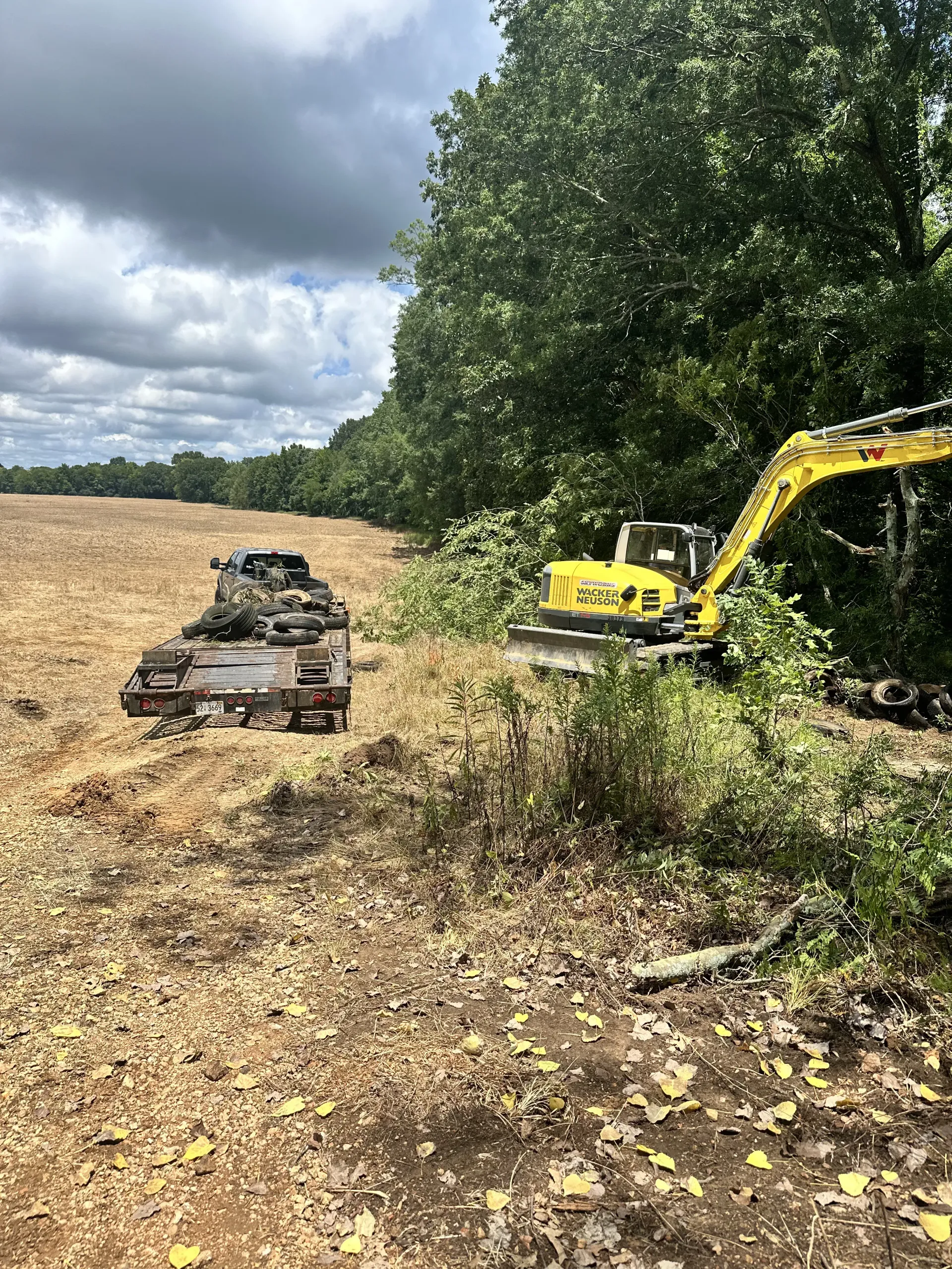 A yellow excavator clears brush at the edge of a field next to a dark-colored truck and trailer.