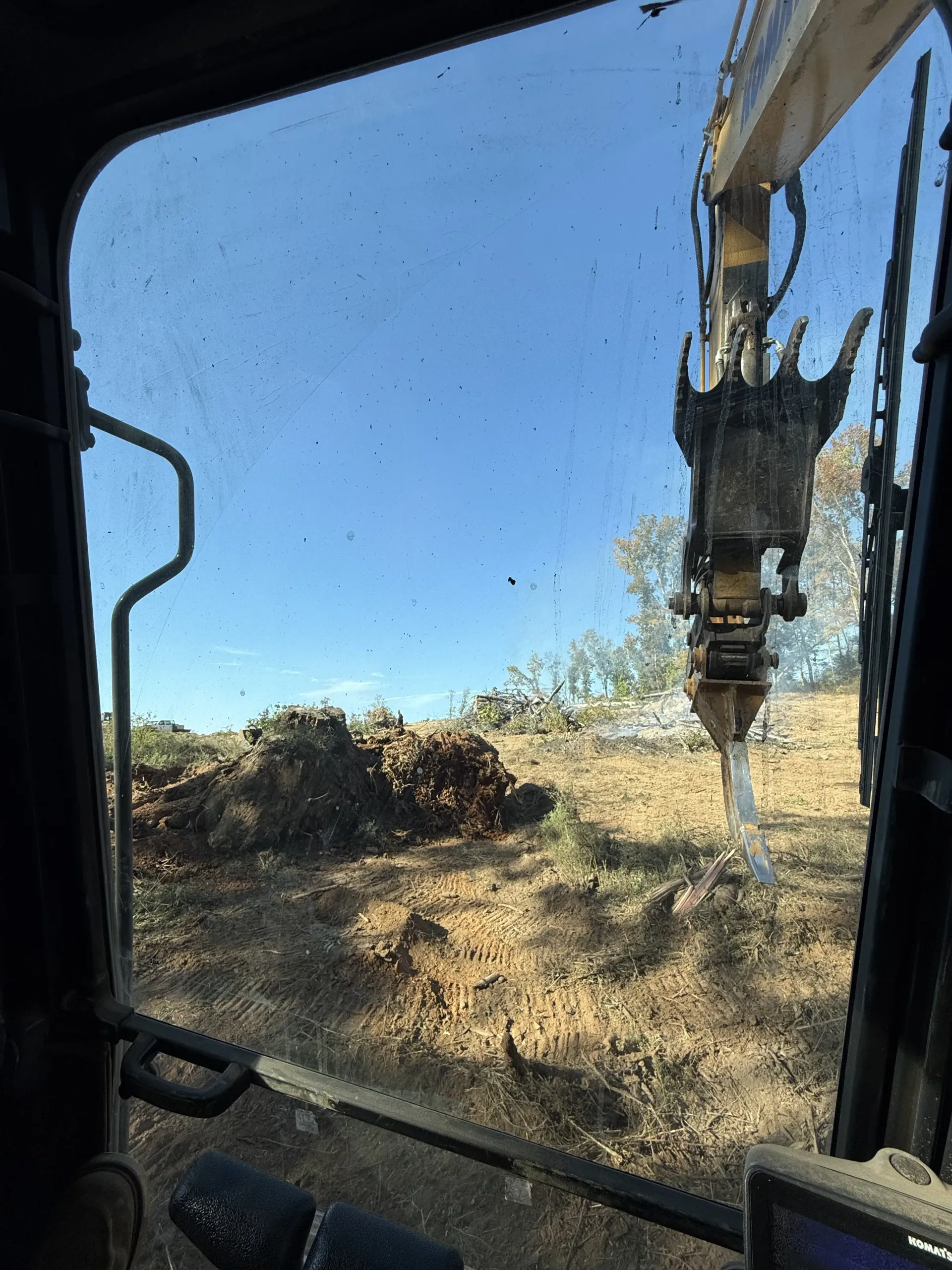 A view from inside an excavator cab looking out at a field of dirt and brush with the machine's metal claw visible.