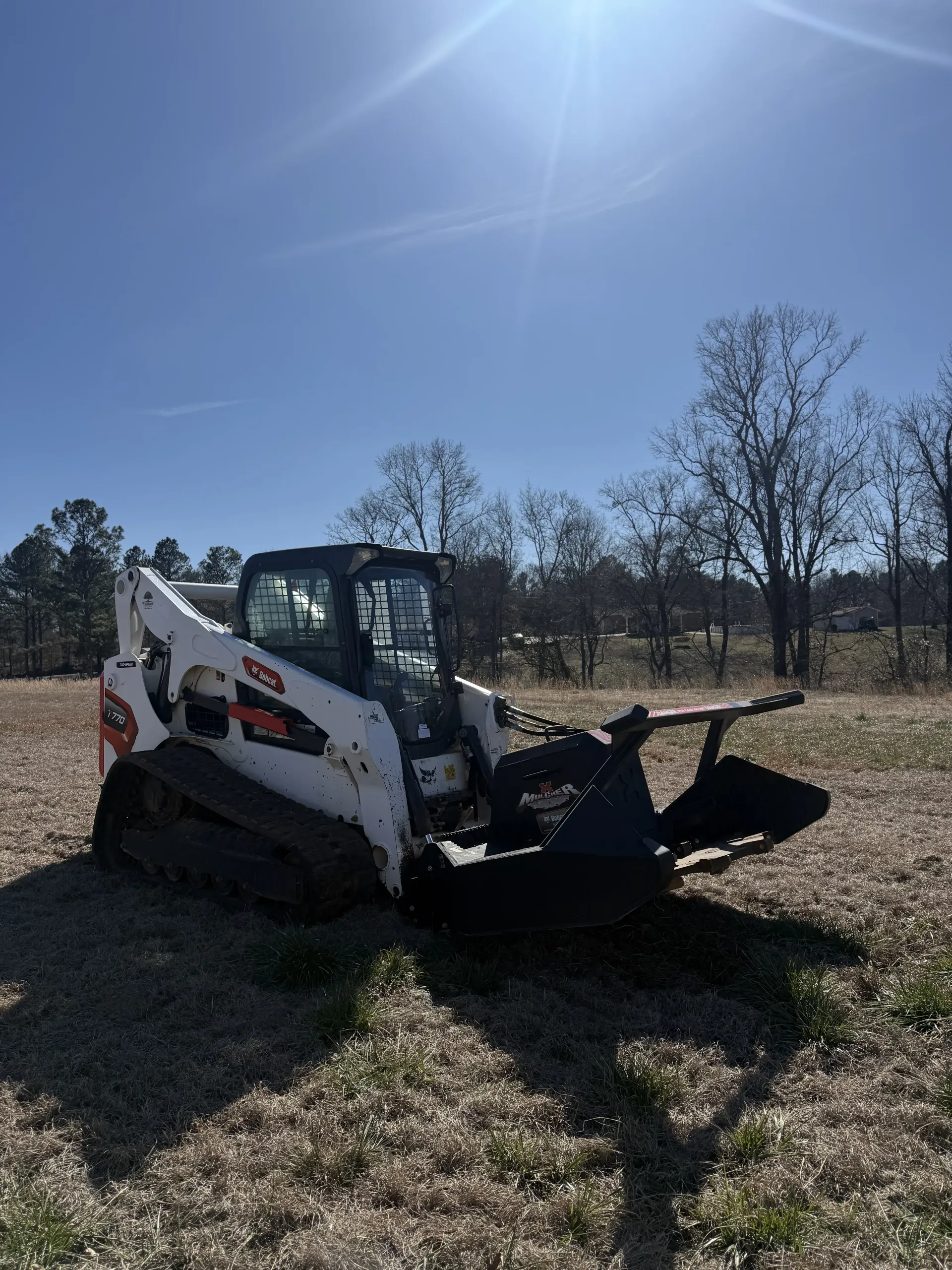 A white Bobcat compact track loader with a mulching attachment sits in a sunny, cleared field with sparse trees.