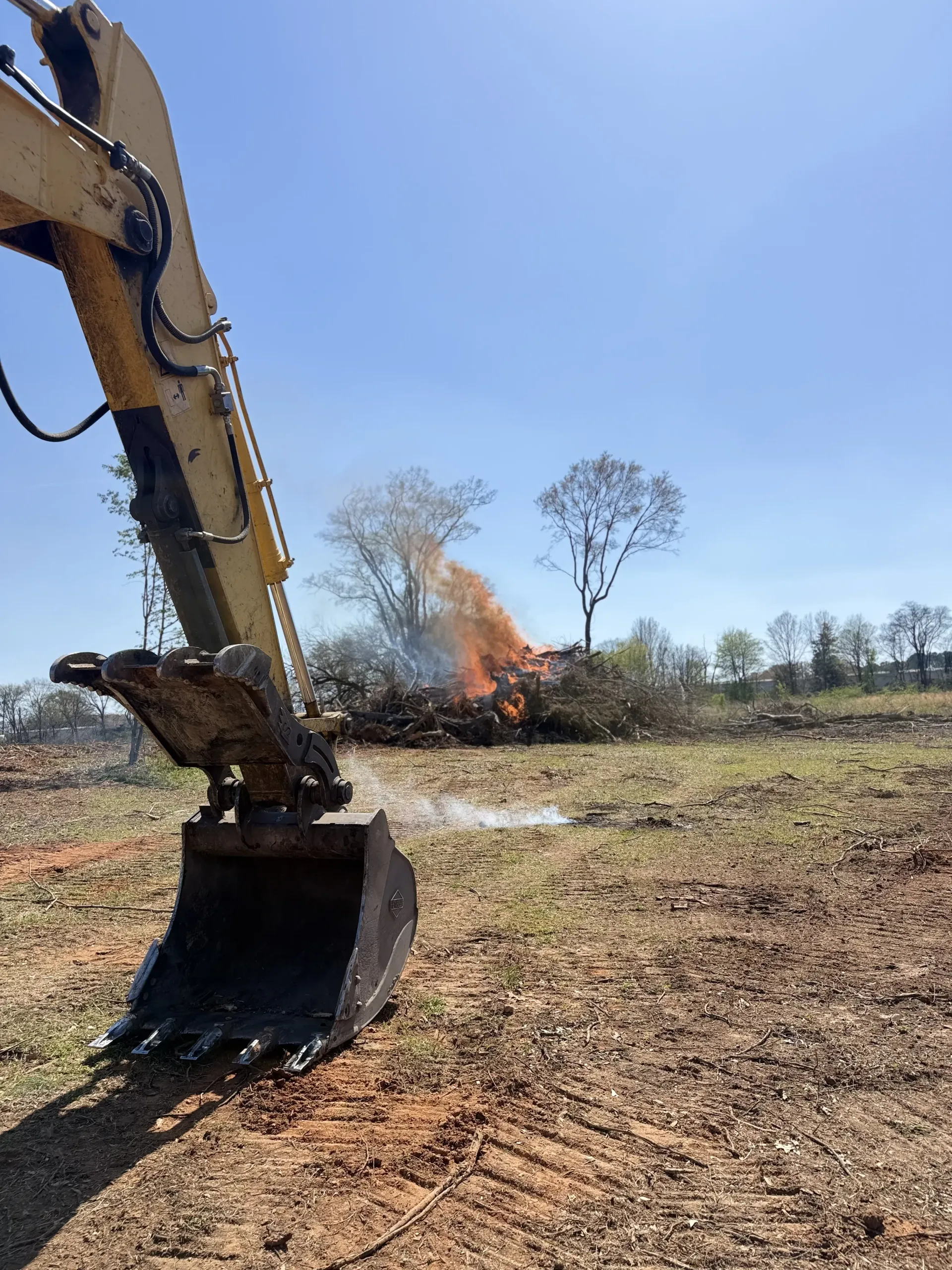 An excavator bucket in the foreground faces a pile of brush burning in a cleared, sunny field.