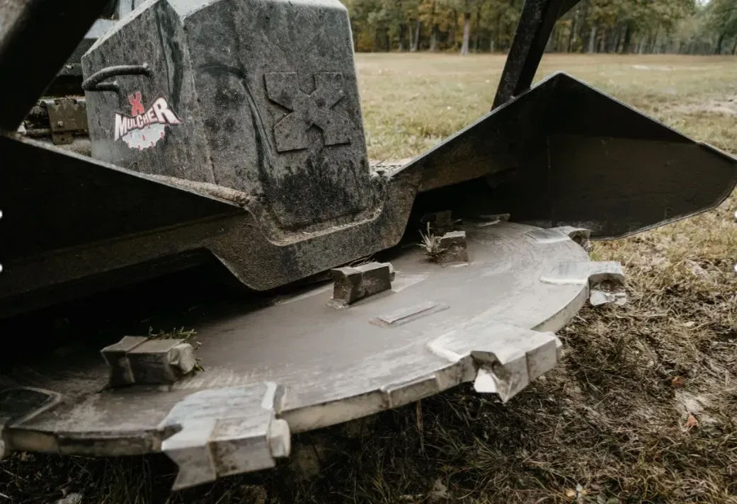 A heavy-duty brush cutter attachment with sharp metal teeth, mounted on machinery in a grassy field.