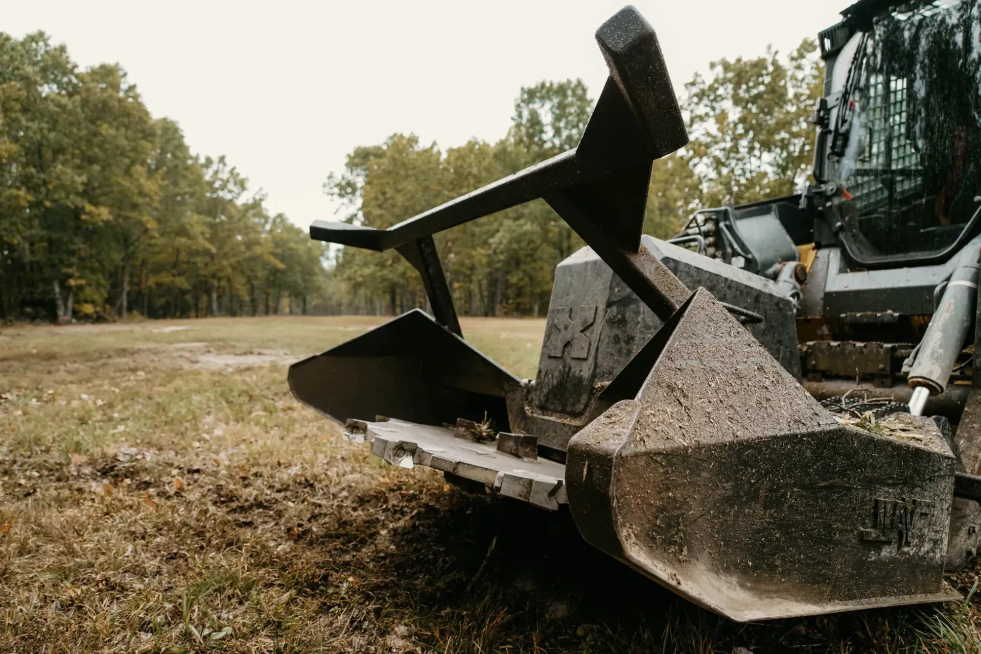 Close-up of a skid steer tree shear attachment in a grassy field with trees in the background.