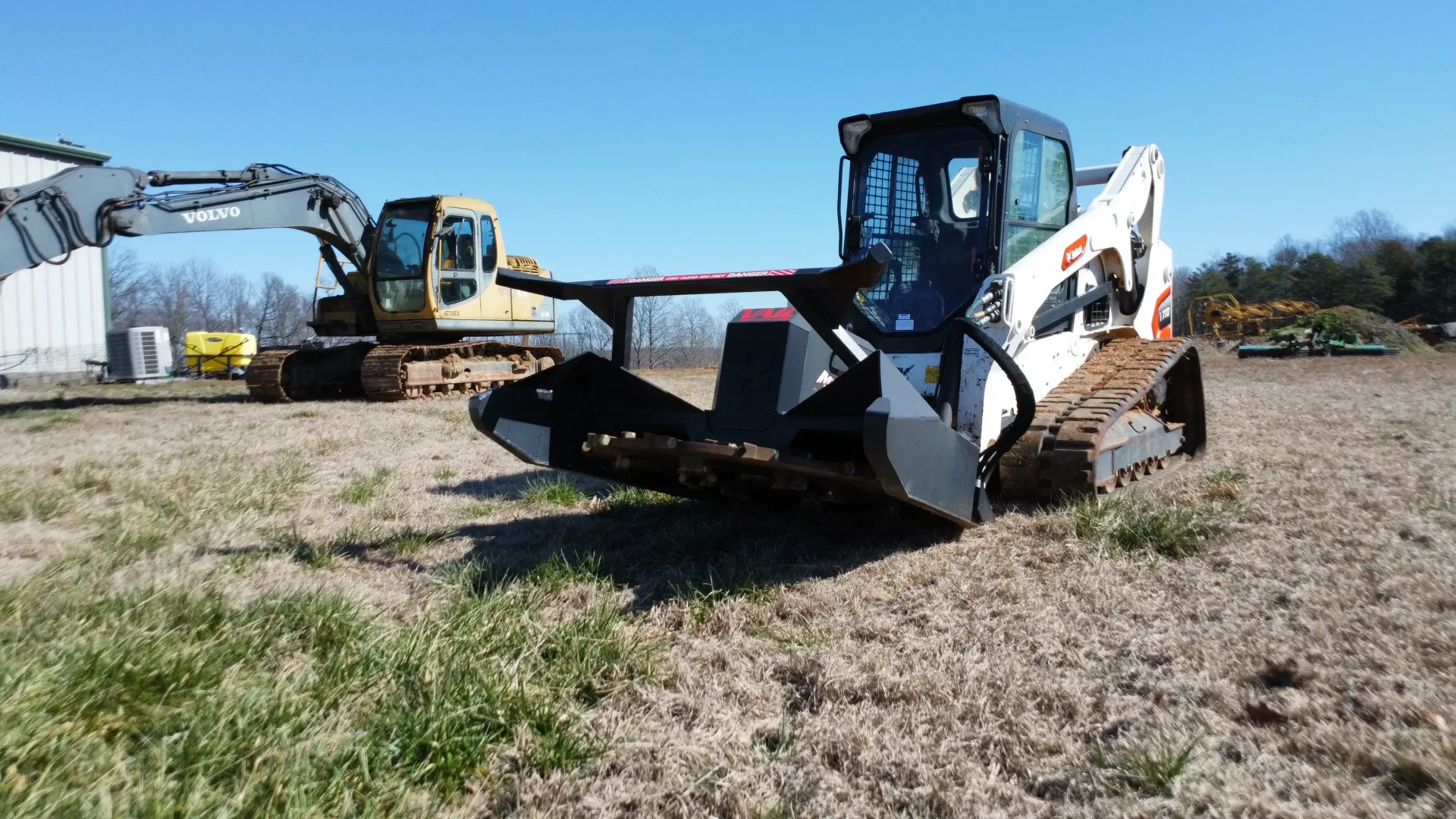 A white Bobcat compact track loader with a metal forestry attachment, parked in a field with an excavator in the background.