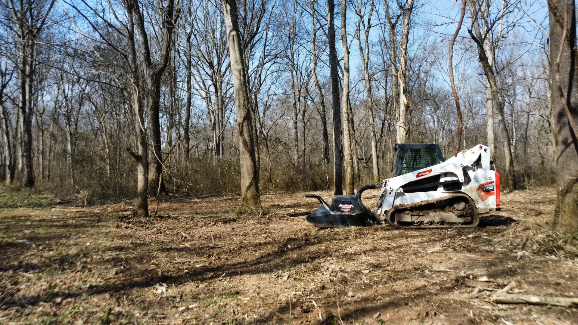 A white compact track loader with a forestry mulcher attachment clearing brush in a wooded area.