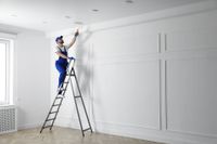 A painter in blue overalls, on a ladder, rolling white paint onto a wall.