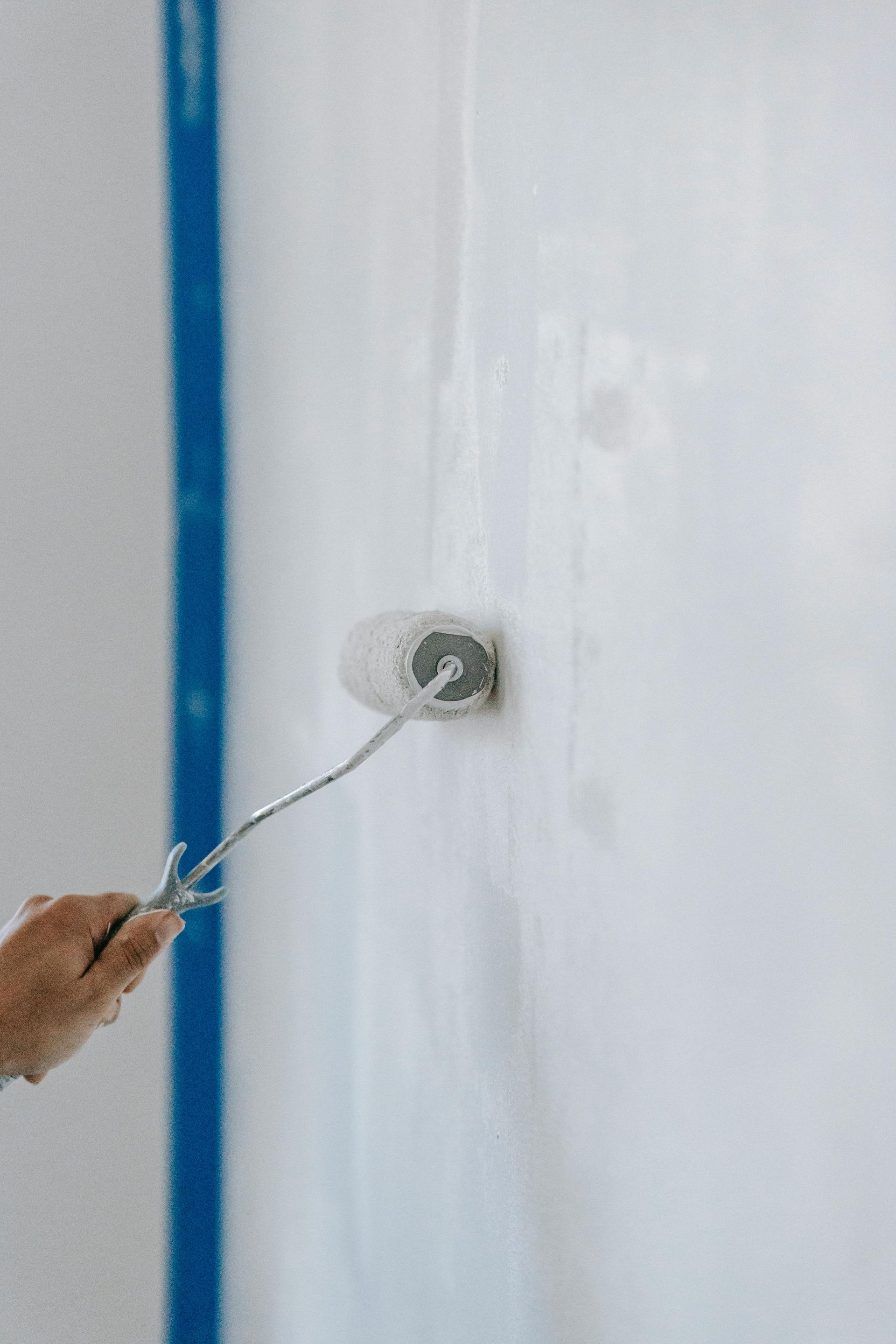 Person painting a white wall with a paint roller, blue tape on the edge.