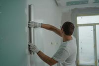Person using a long, metal tool to smooth drywall on a wall.