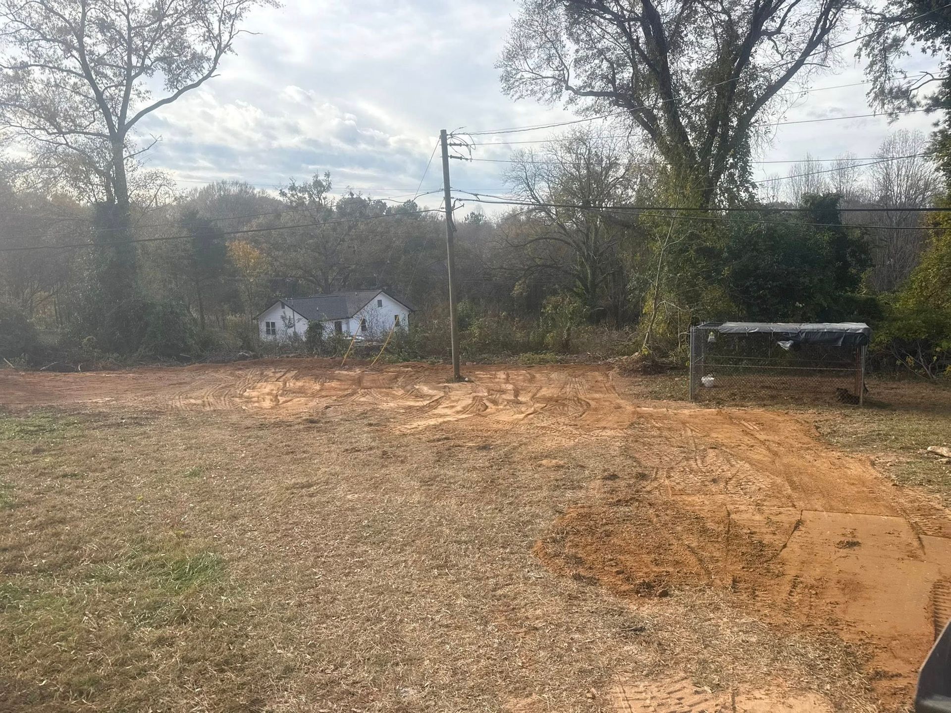 A cleared lot with a dirt foreground, small white houses in the distance, and trees overhead.