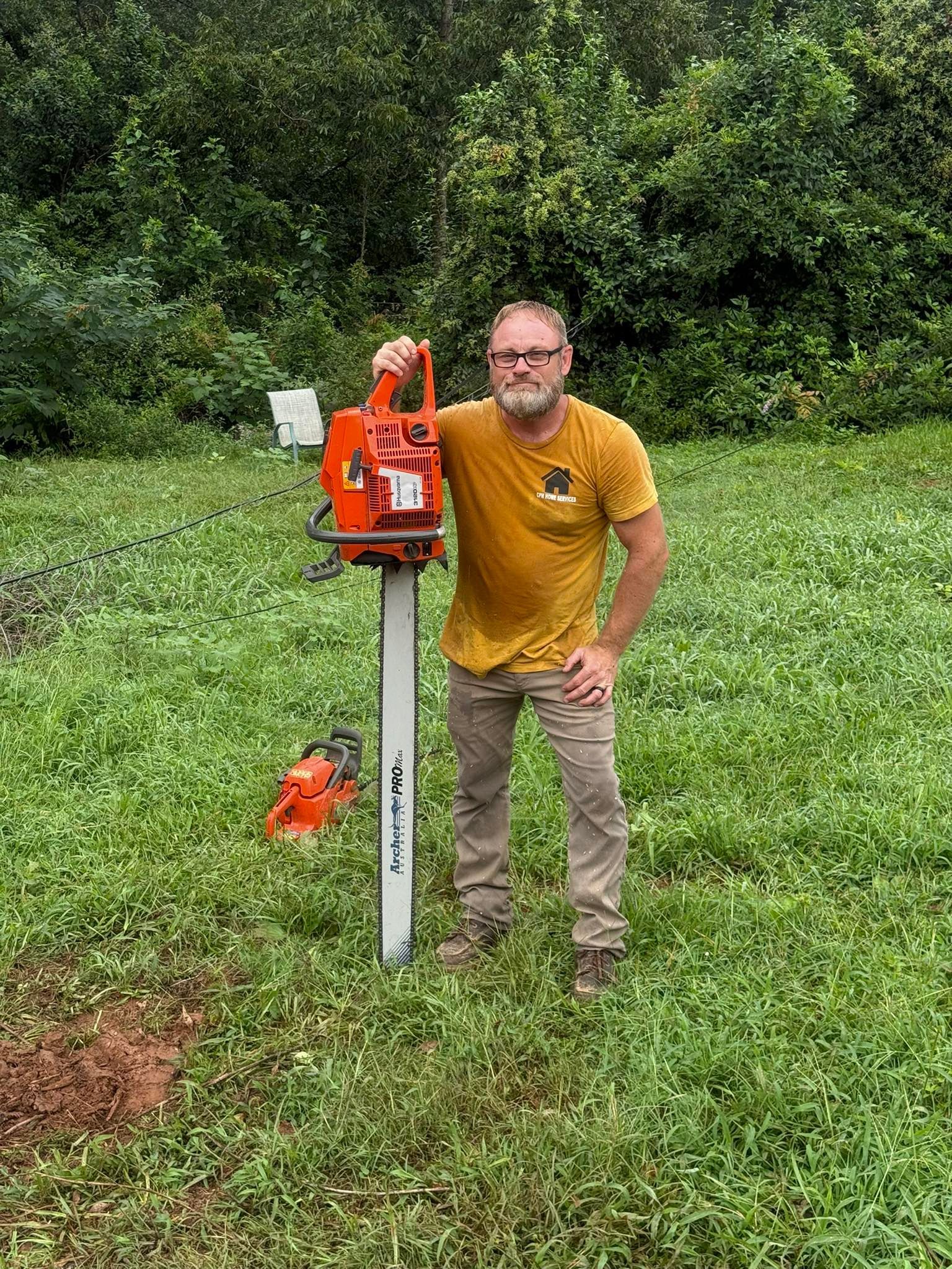 Man holding an orange chainsaw outdoors. Second chainsaw on the ground. Green grass and trees in background.