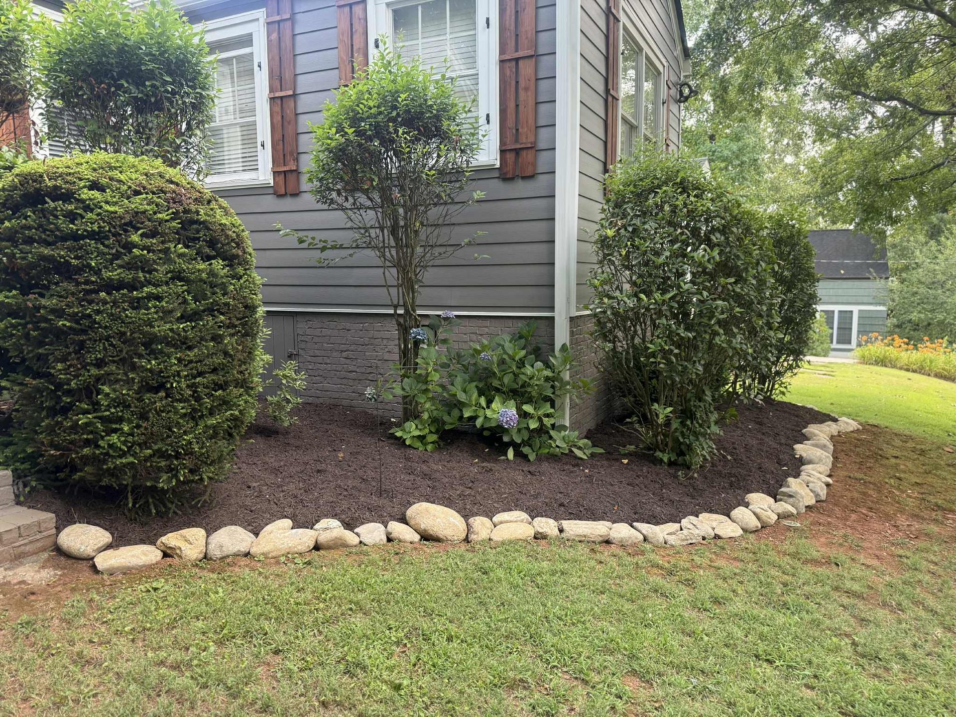 Corner of a gray house with a landscaped bed. Green bushes and mulch border edged by rocks.