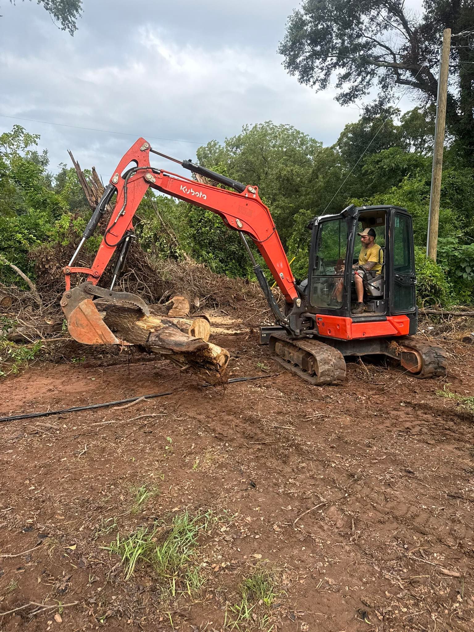 Orange Kubota excavator clearing debris from a muddy area with a person in the cab. Green trees are in the background.