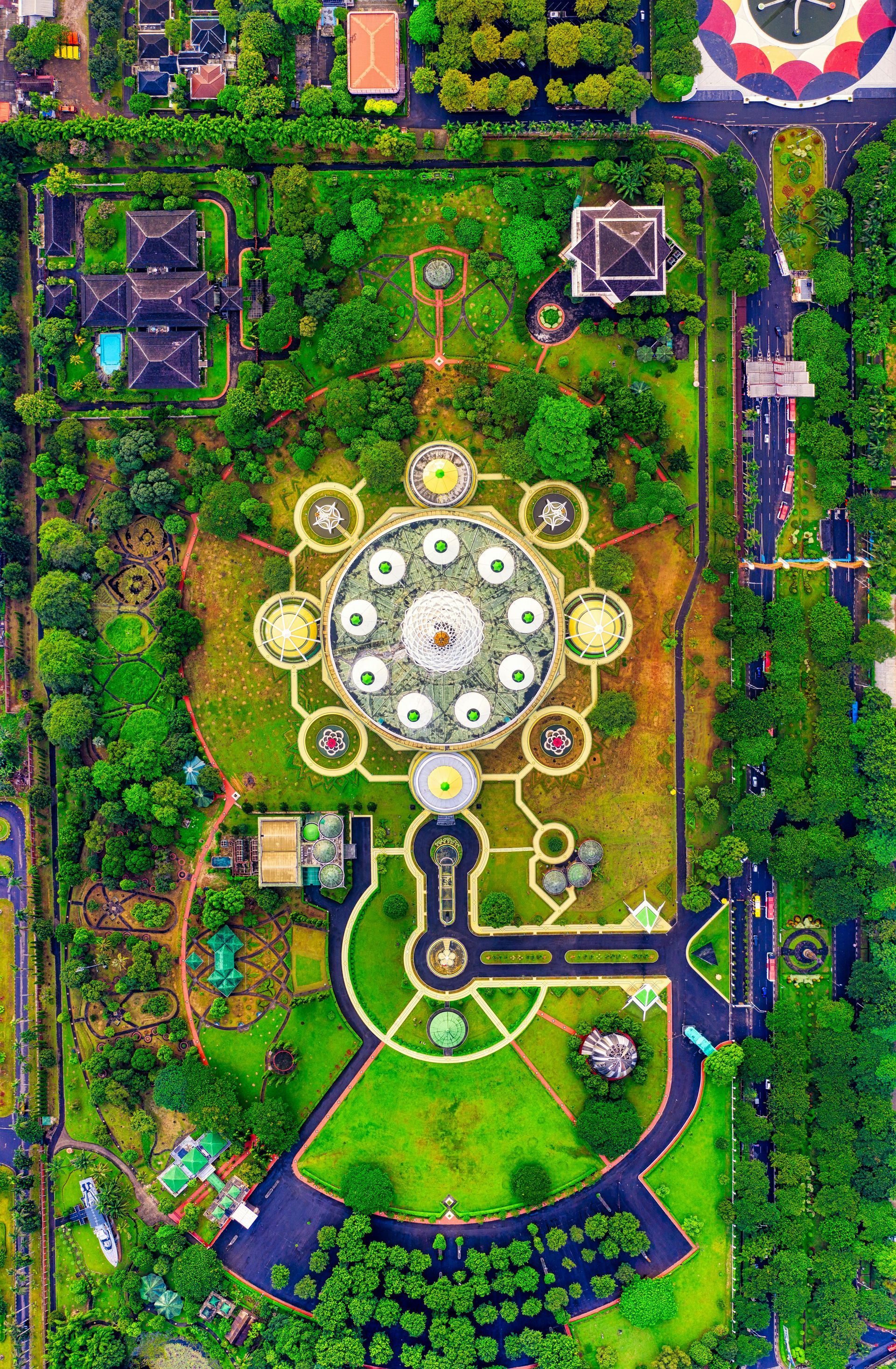Aerial view of a circular, ornate structure surrounded by green lawns and trees. Paved pathways lead to the building.