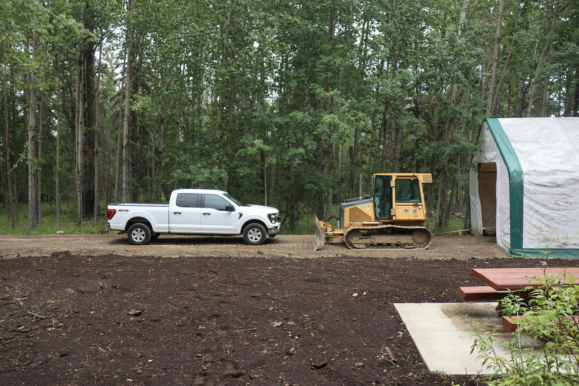 White pickup truck and bulldozer on dirt near a tarp-covered building, trees in the background.