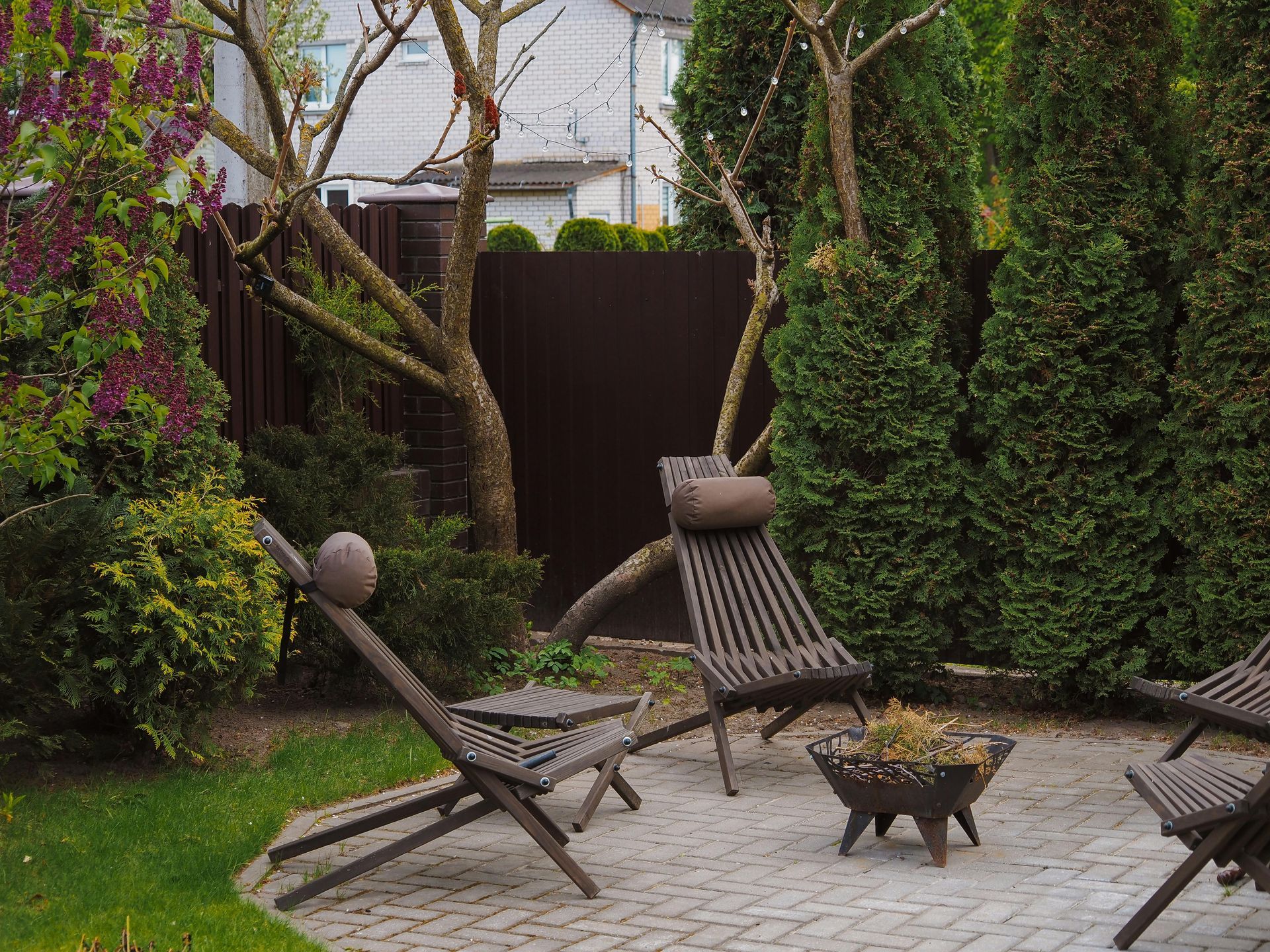 Patio with wooden chairs, small table, fire pit, and various plants against a brown fence.