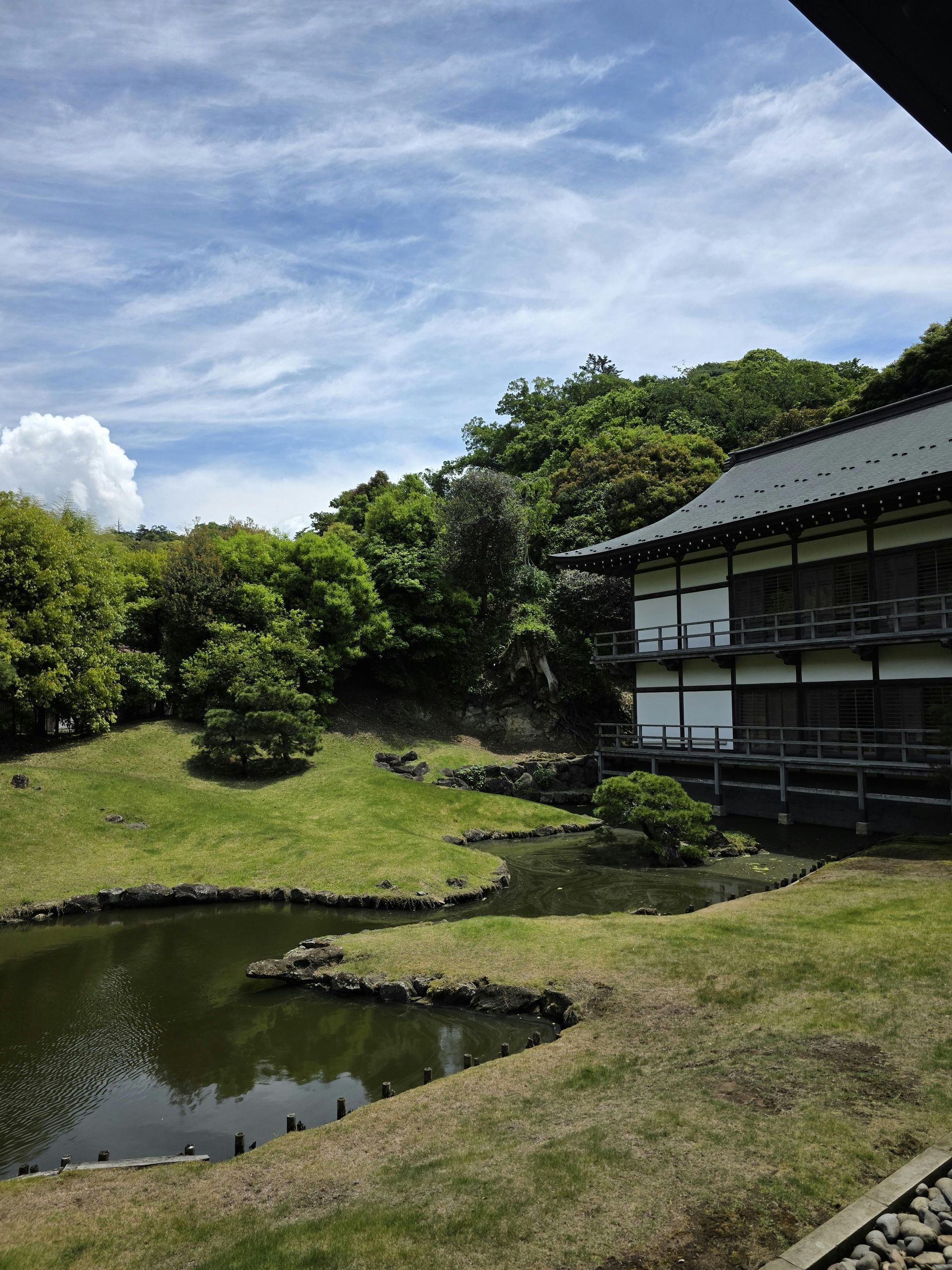 Japanese garden with pond, lush greenery, traditional building, and blue sky.