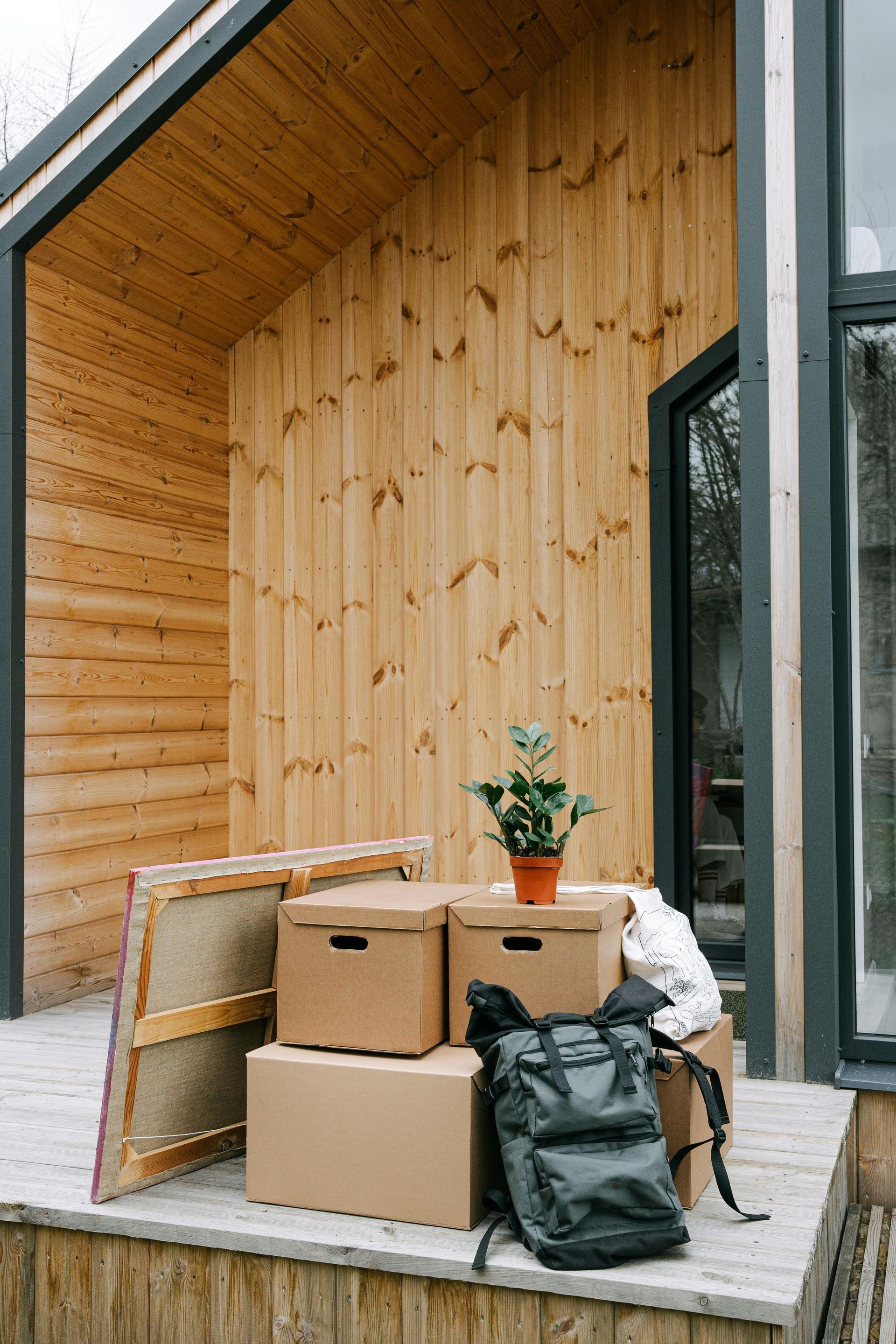 Boxes, backpack, and plant on a porch of a wooden cabin, suggesting a move.