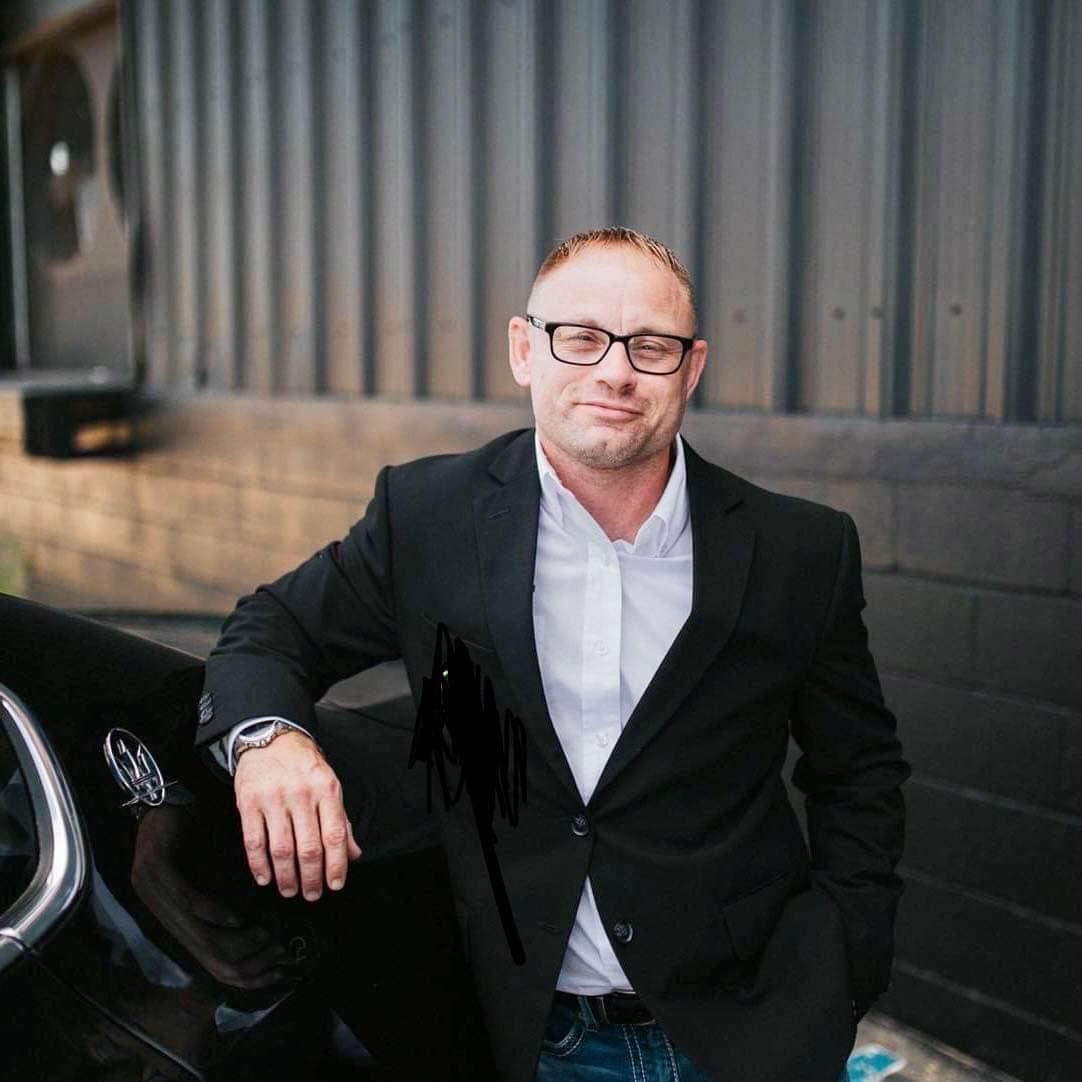 Man in a suit, leaning on a car, wearing glasses, smiling. Brick wall background.