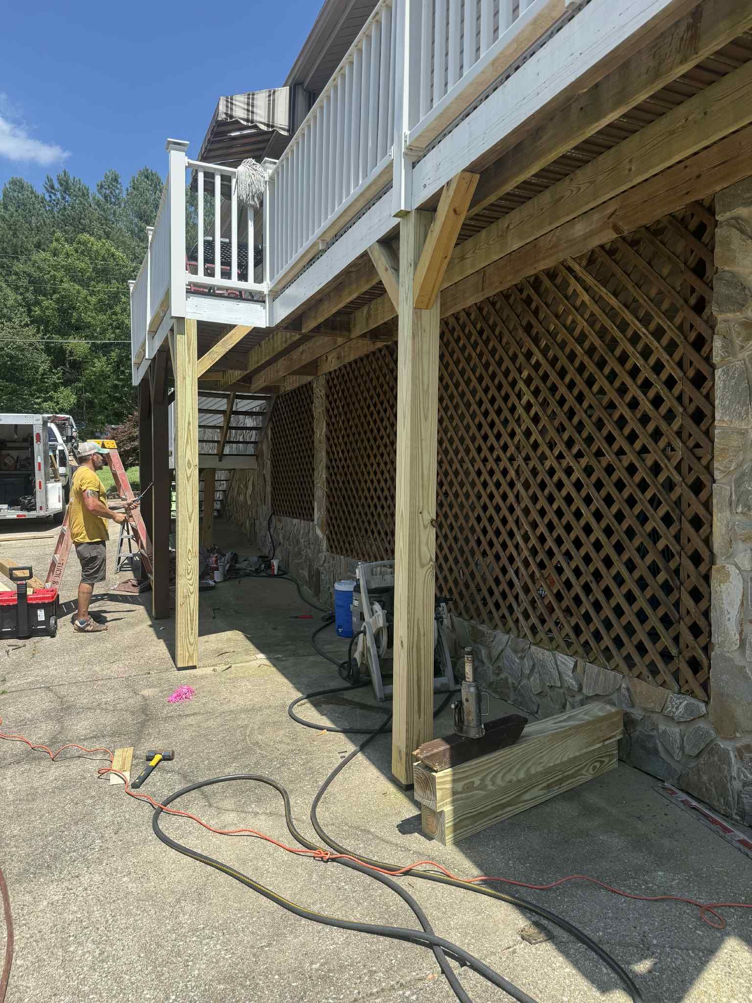 Deck construction: A person works on wooden supports under a deck with lattice siding, on a stone foundation.