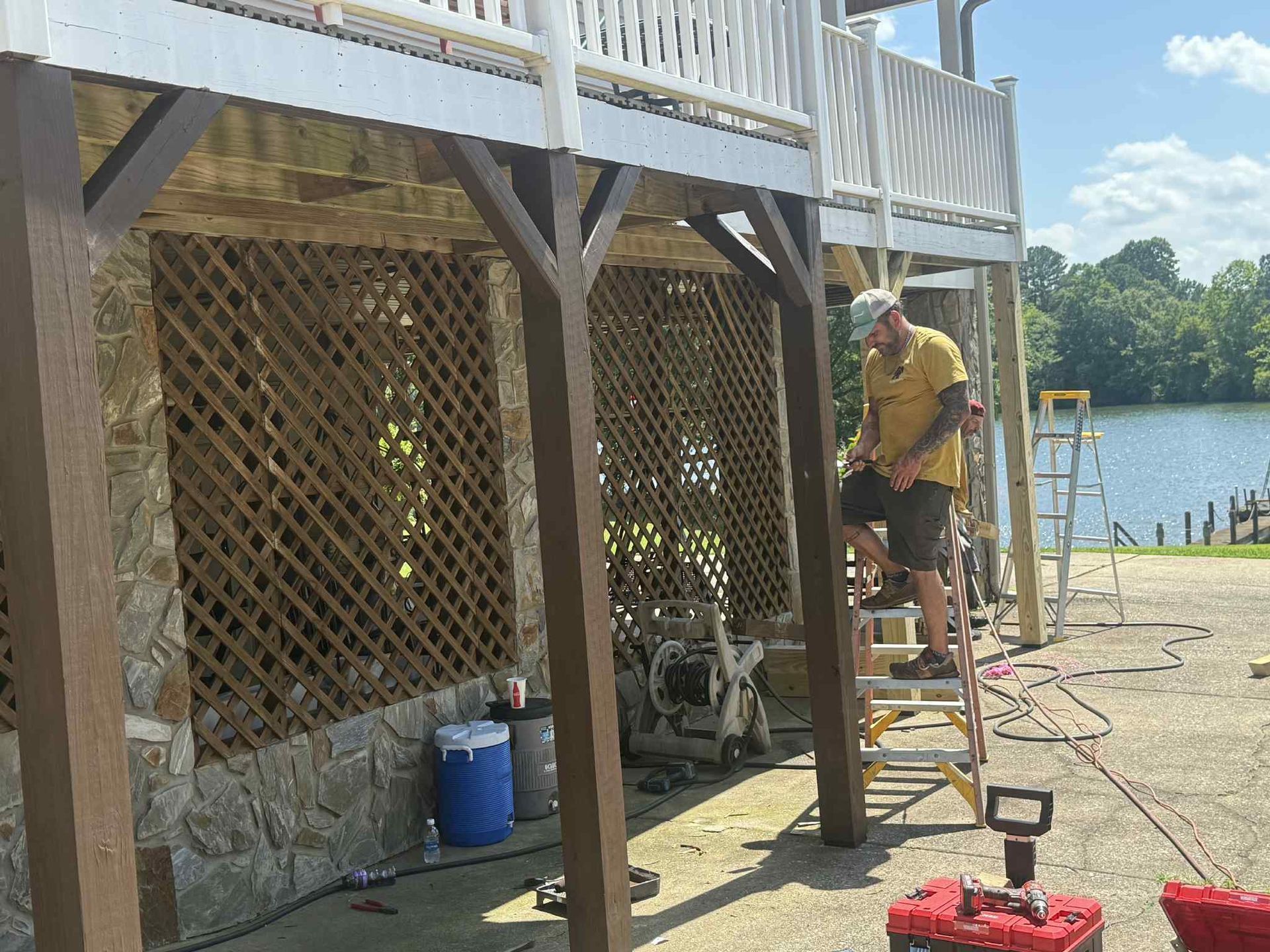 Man on ladder working on a deck. Brown columns, lattice, and blue cooler. Lake visible in the background.