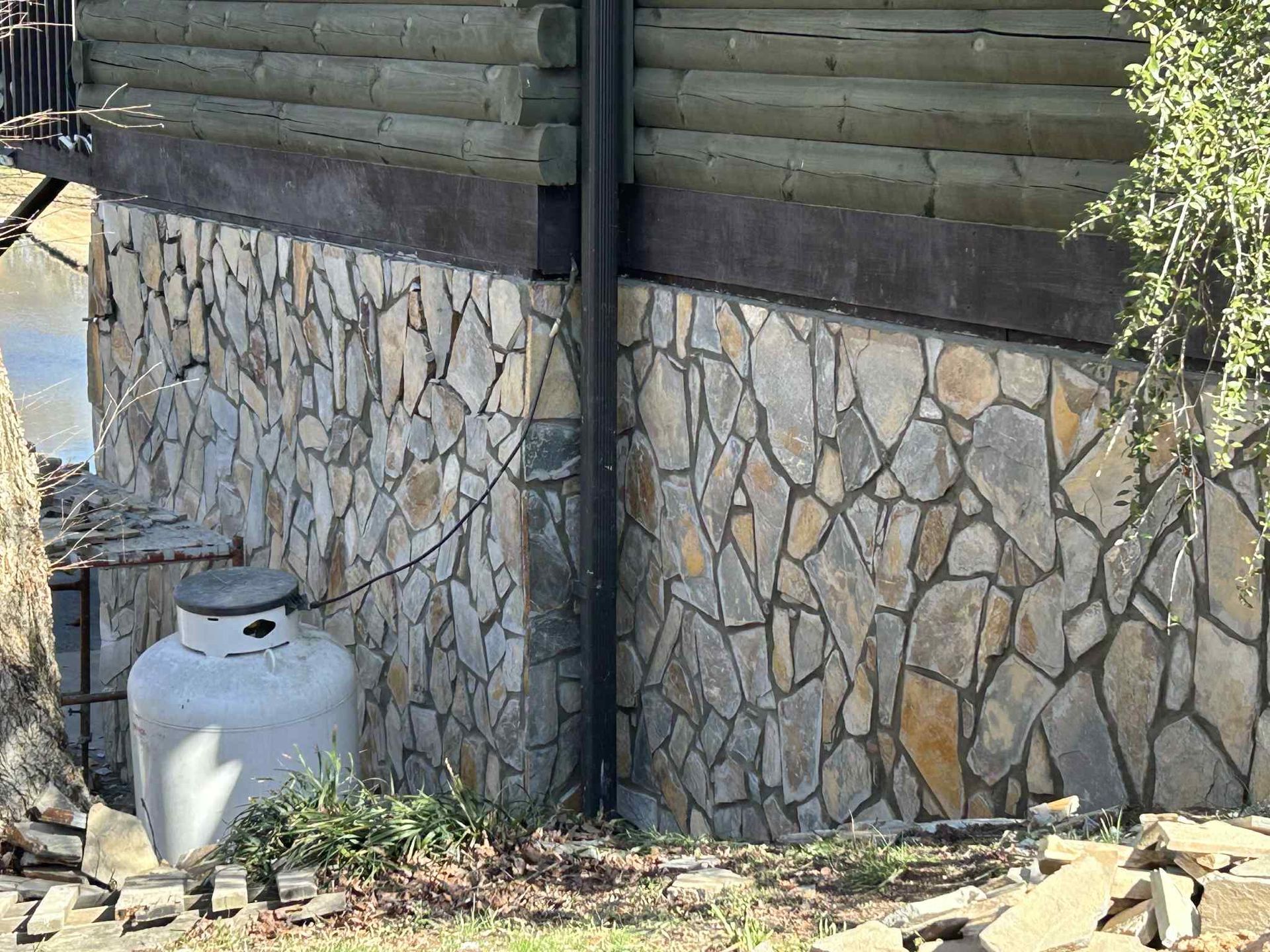 Stone facade of a building with log siding above. A propane tank sits on the ground nearby.