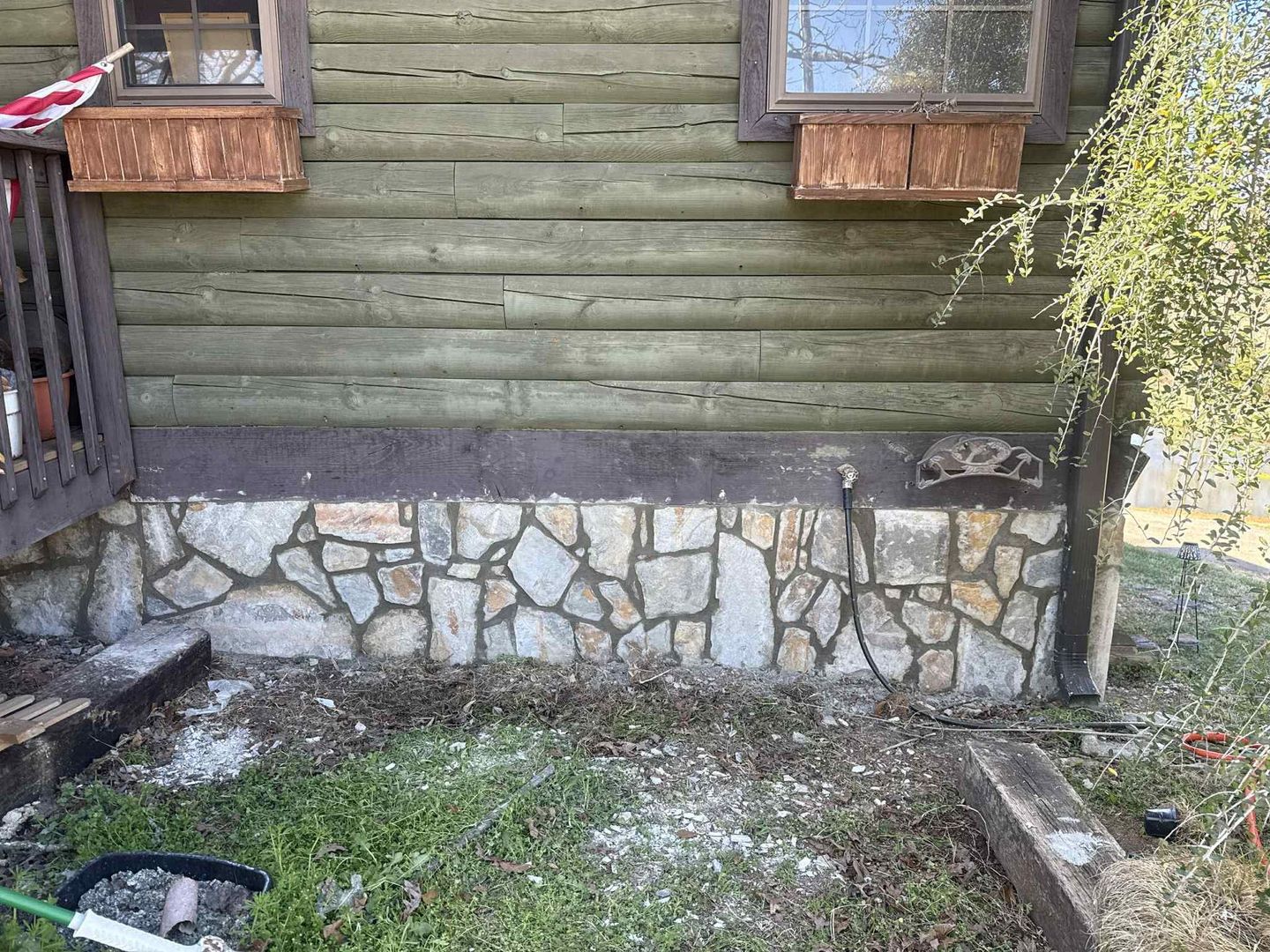 Stone foundation with green-painted logs above, two windows with flower boxes. Yard debris around base.