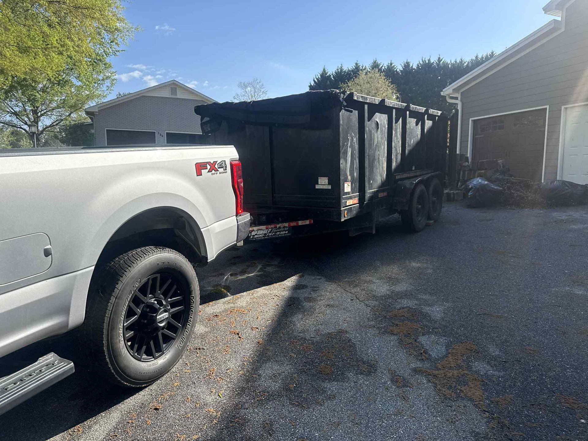 White truck towing a black dump trailer on a gravel driveway next to a house.