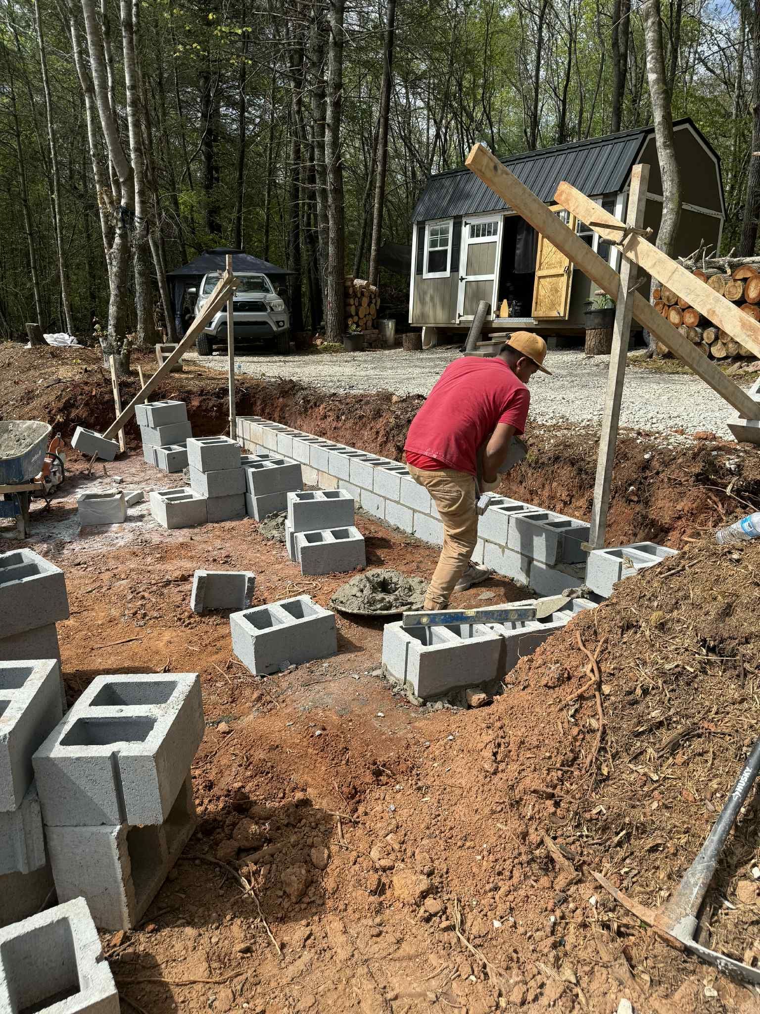 Man laying cinder blocks for a foundation in a wooded area. Tiny house in the background.