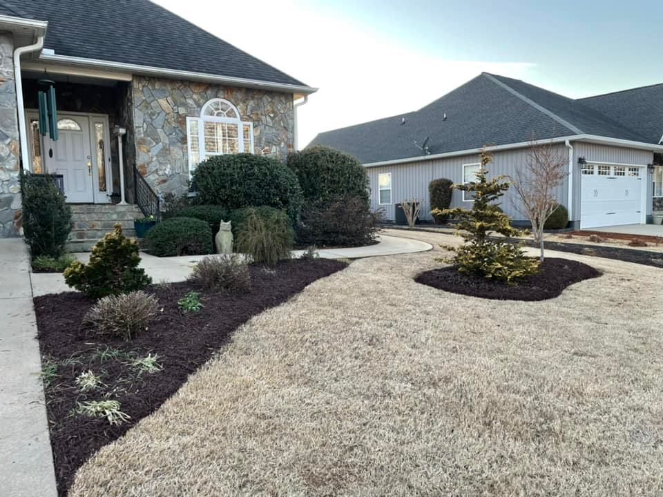 Front yard of a stone house with brown mulch beds, green shrubs, and dry grass. A detached garage is in the background.