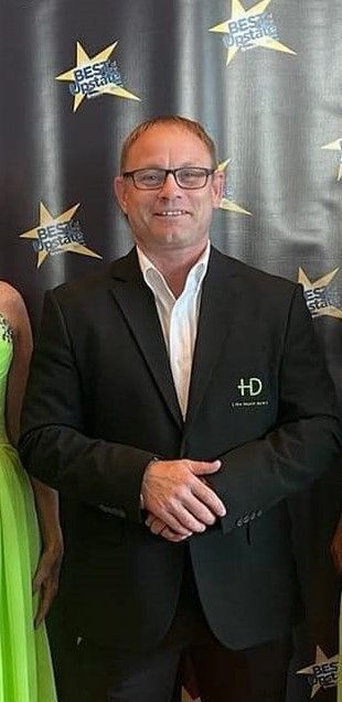 Man in black suit and glasses, arms crossed, standing in front of a backdrop with star decorations.