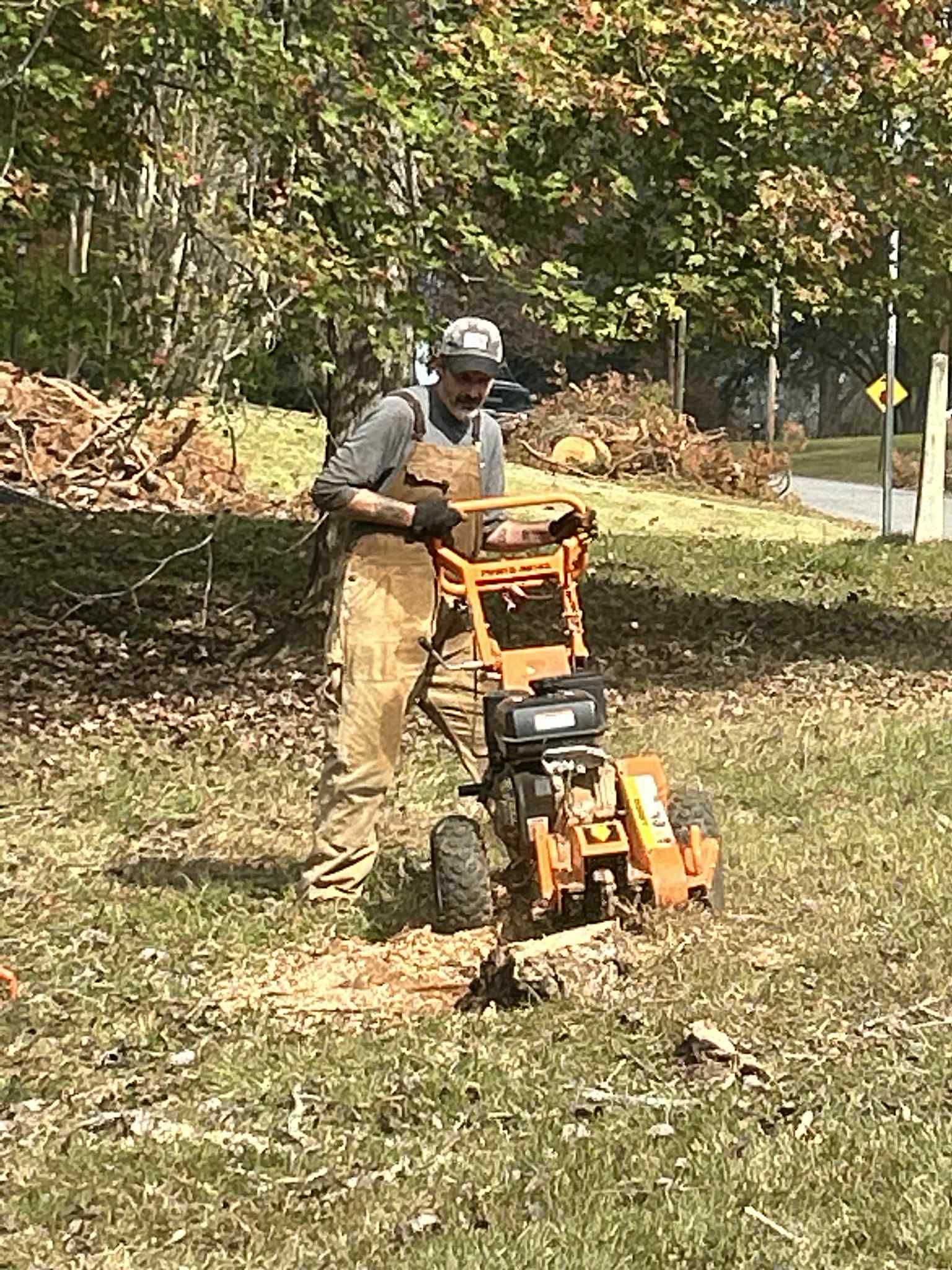 Man operating a stump grinder outdoors in grassy area; wood chips visible.