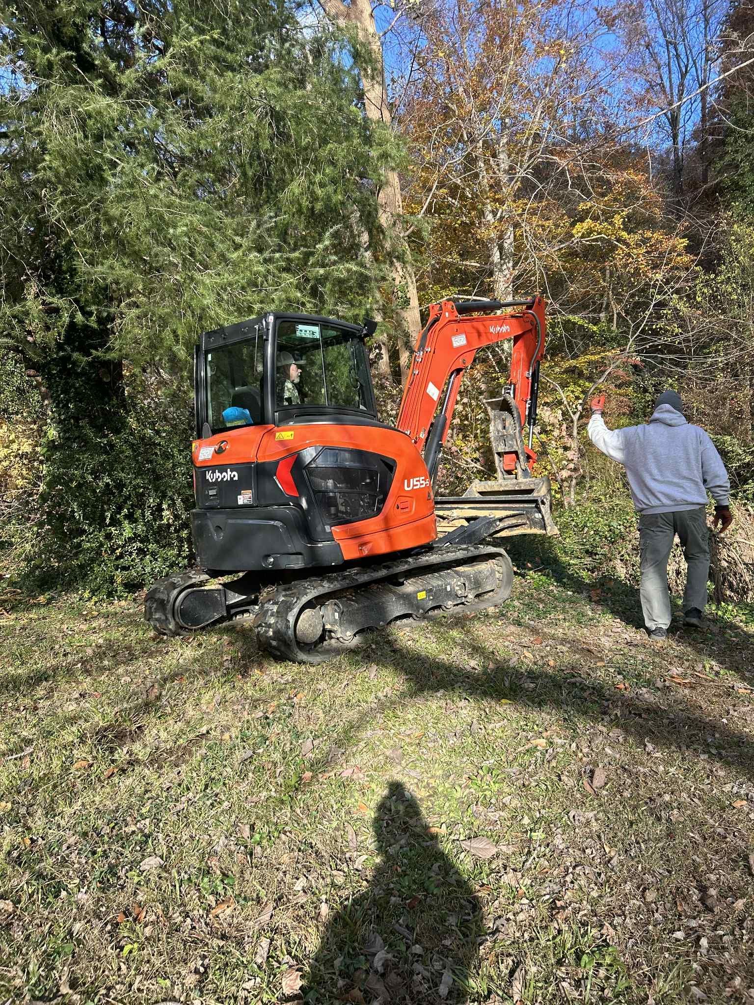 Orange excavator in a wooded area; person waves nearby.
