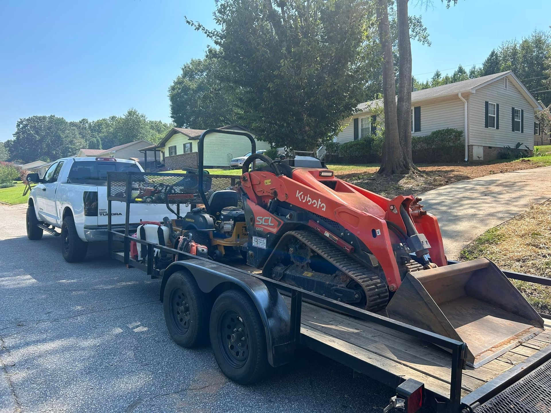A white pickup truck towing a trailer with landscaping equipment, in a residential area.