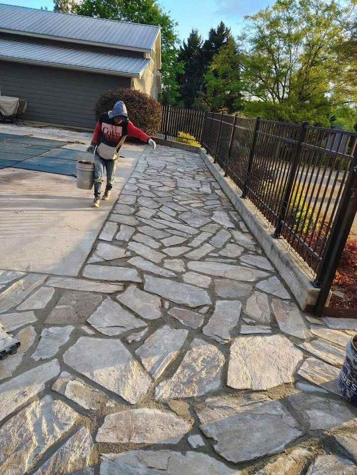 Person walking on stone path, holding bucket, near a black fence and building.