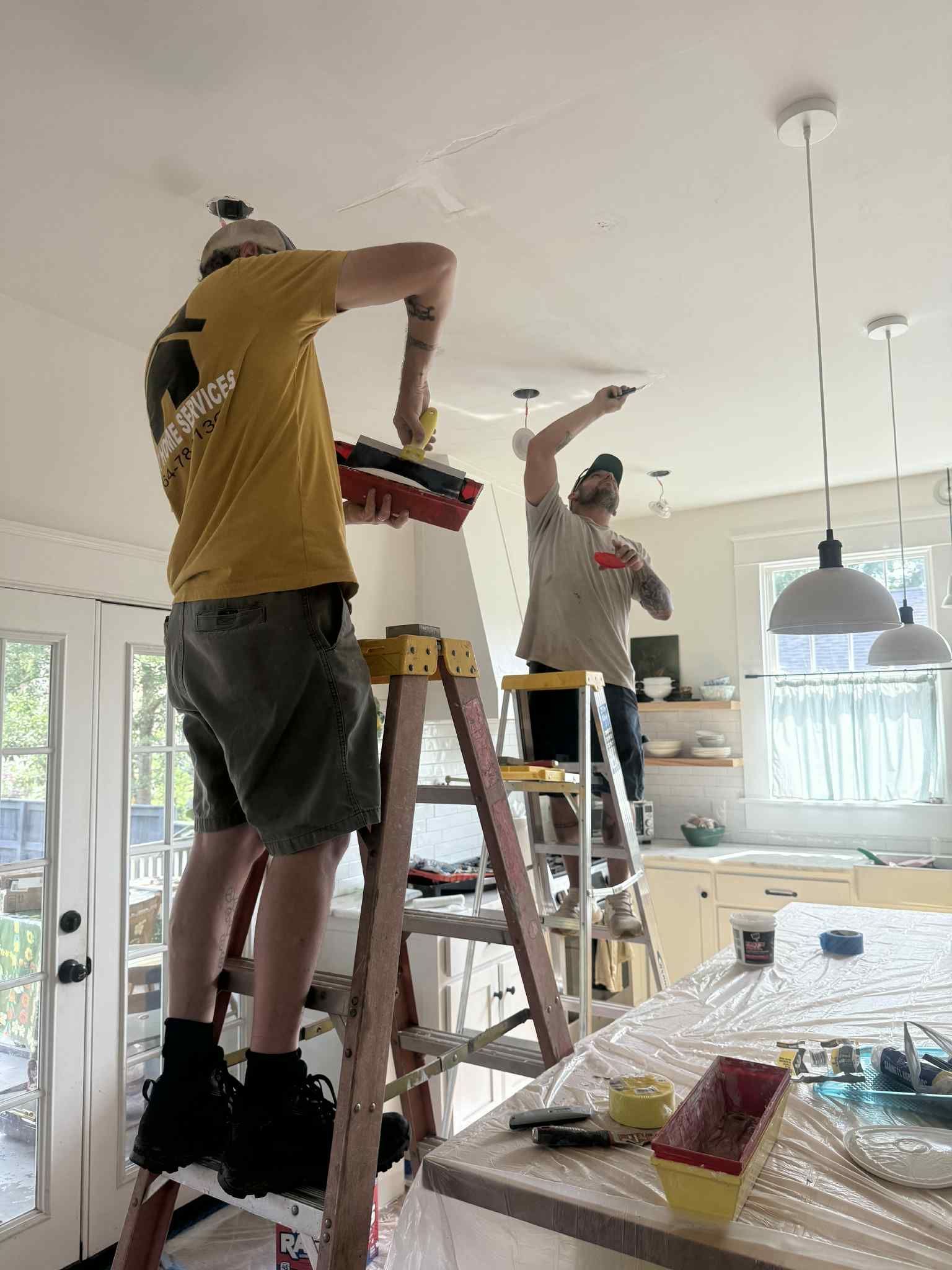 Two people painting a ceiling, one on a ladder, in a kitchen with white walls and pendant lights.