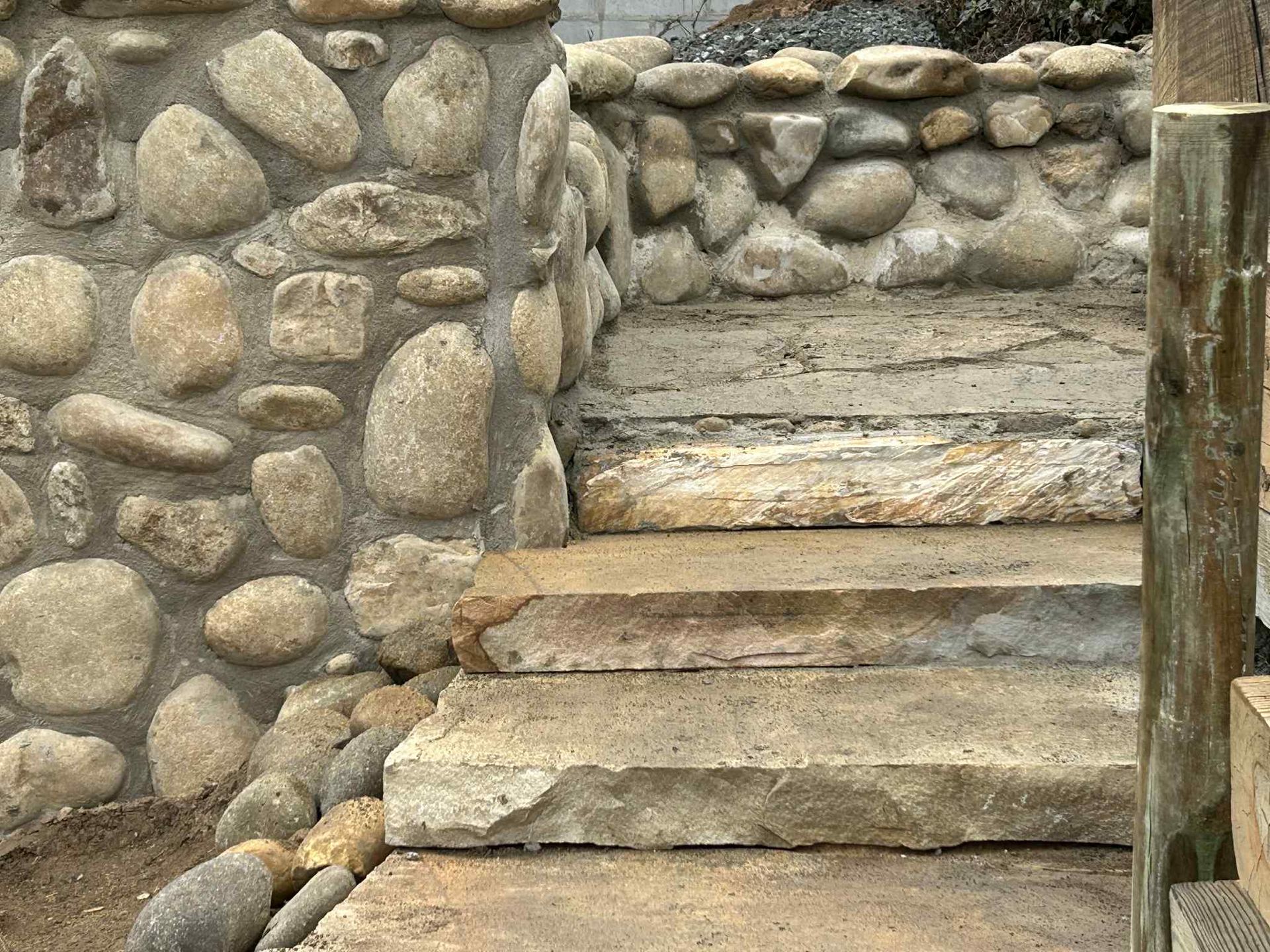 Stone steps next to a stone wall with a wooden post, outdoors.