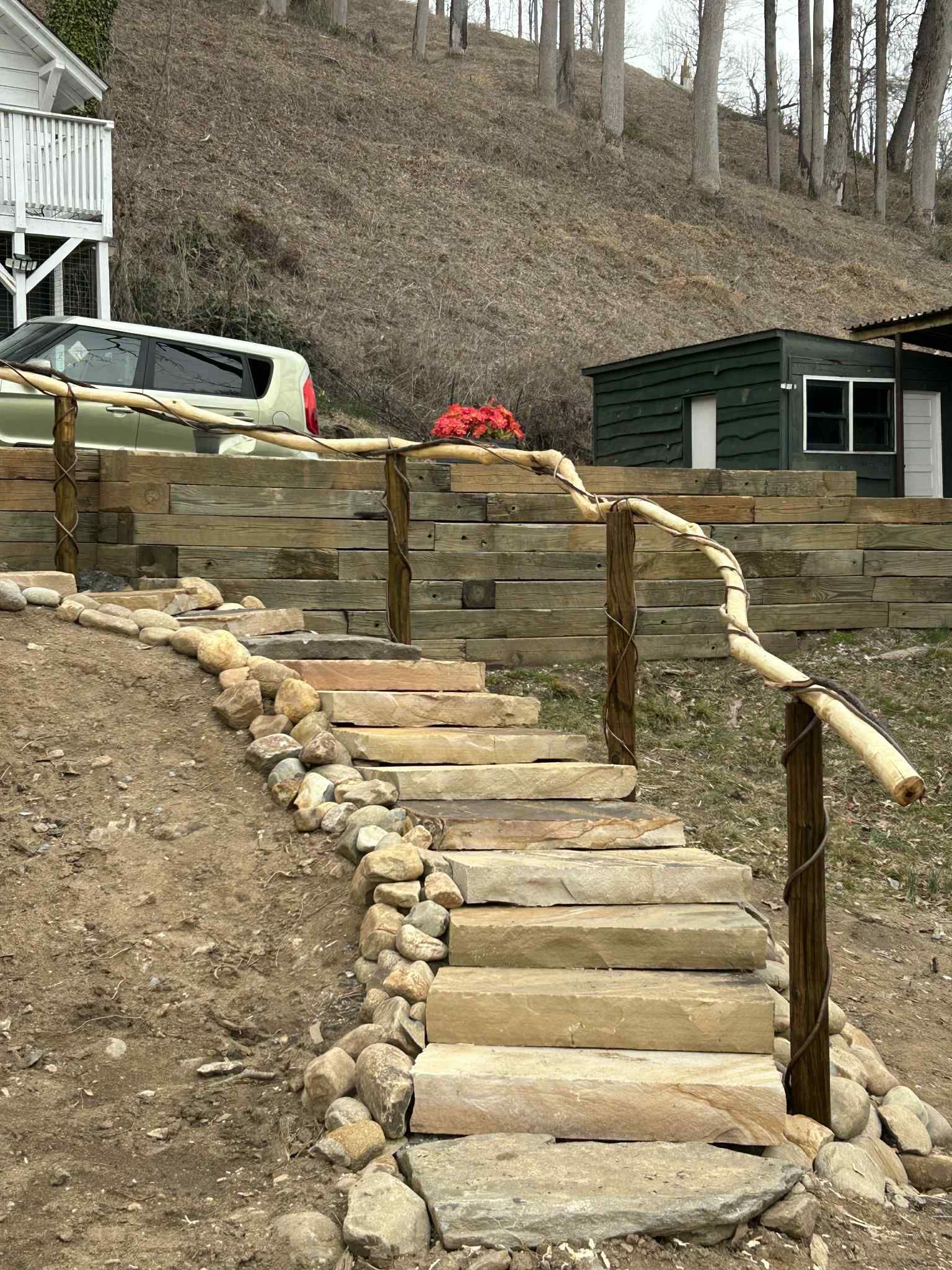 Stone steps with a wooden railing leading up a hillside. Retaining wall and greenery in the background.
