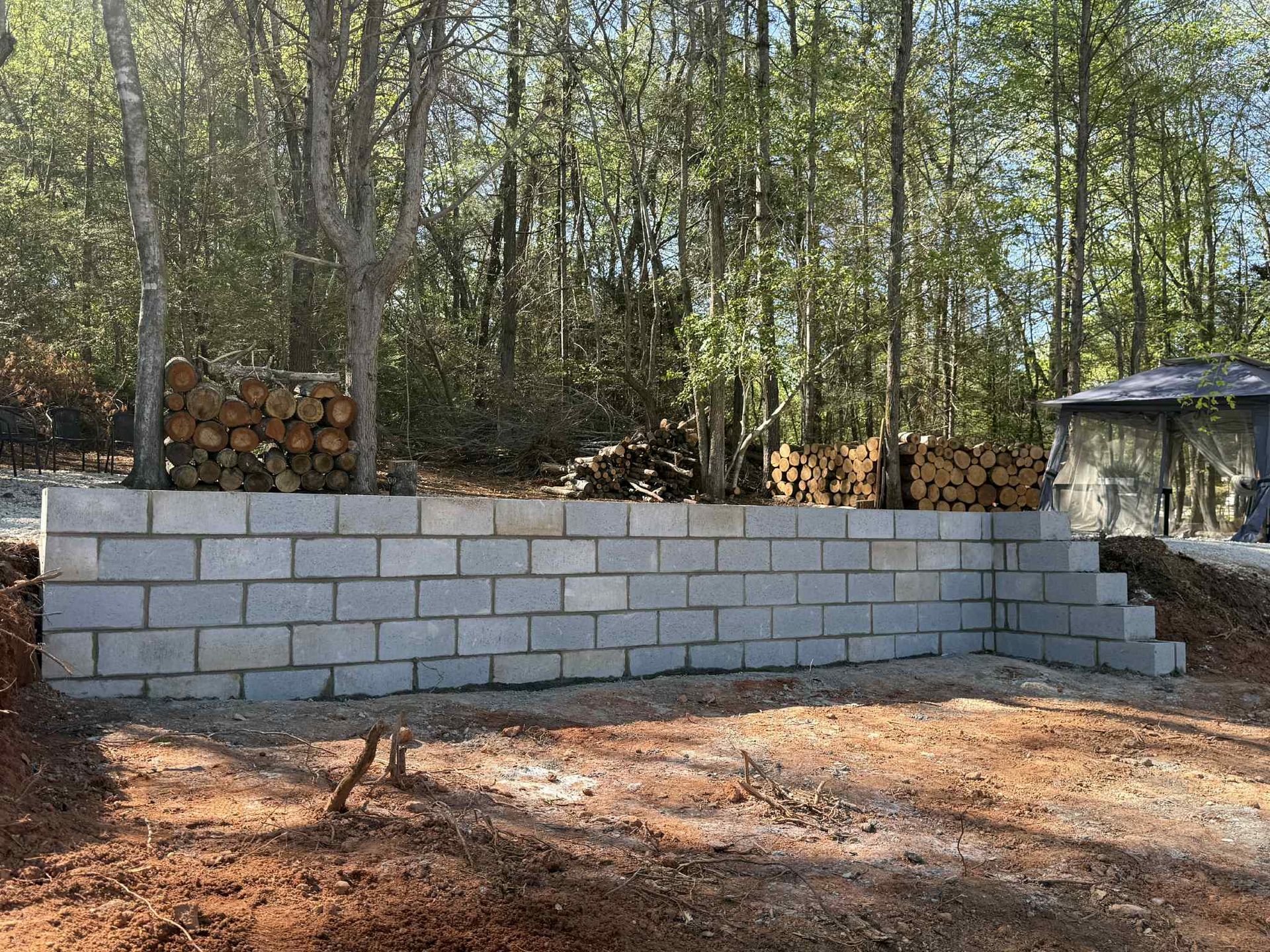 A gray cinder block retaining wall in a yard, with firewood stacked behind it and trees in the background.