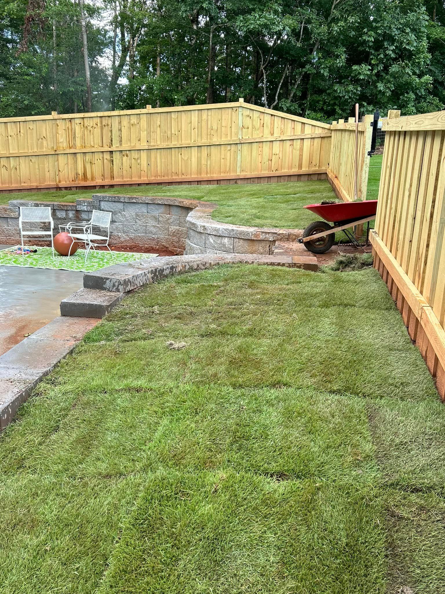 Newly laid green sod in a backyard, beside a stone wall and wooden fence; red wheelbarrow.