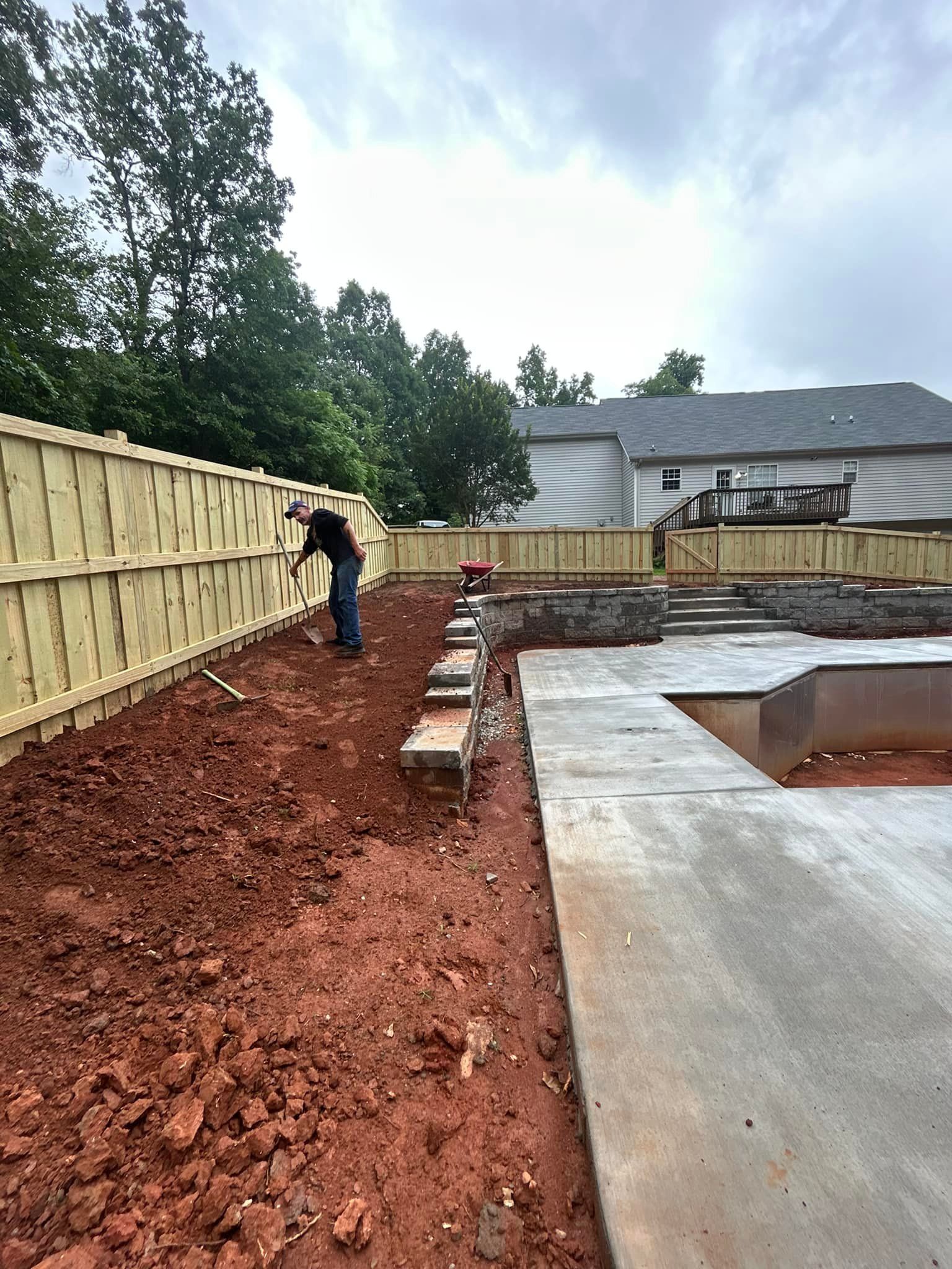 A person shovels dirt next to a wooden fence and a concrete patio under a cloudy sky.