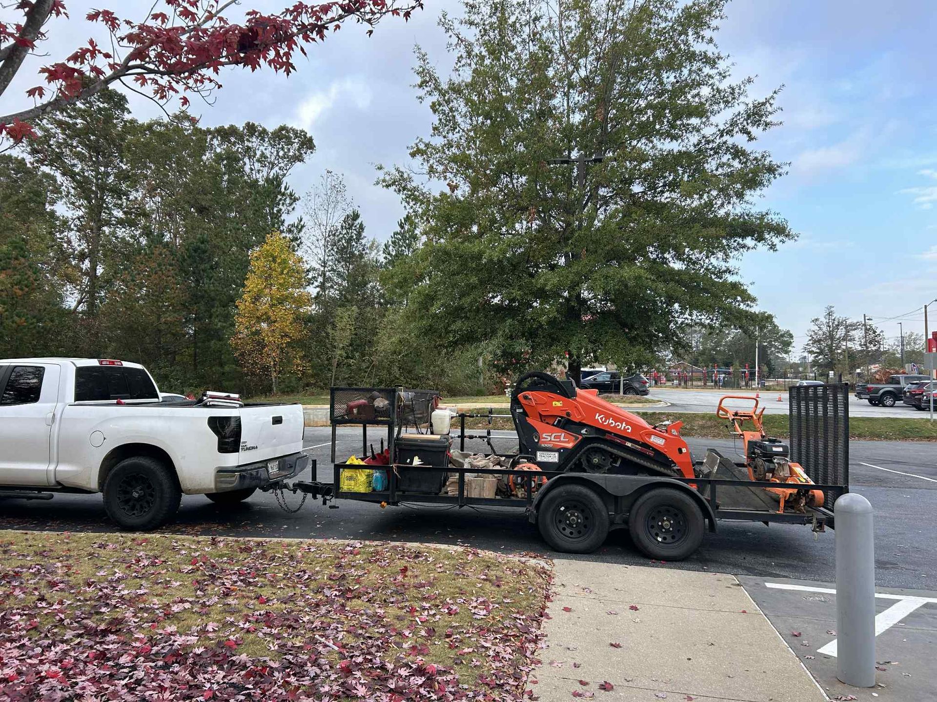 White truck pulling trailer with orange mini-excavator and equipment in a parking lot.