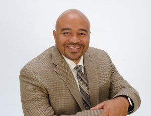 Man in a brown patterned blazer smiles; light background.