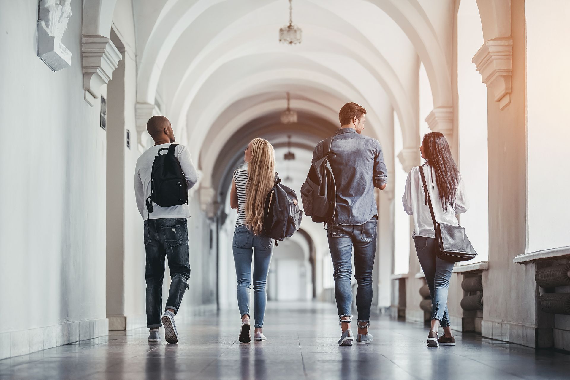 Four students walking down a sunlit arched hallway, carrying bags.