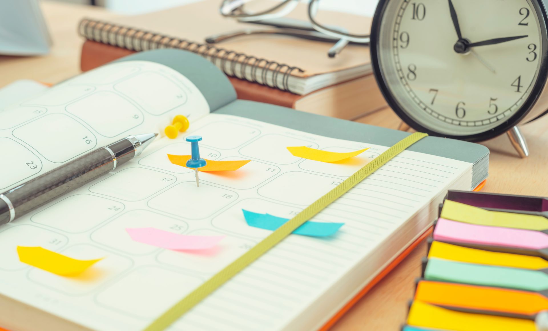 Open planner with colorful sticky notes, a pen, clock, and other office supplies on a wooden desk.