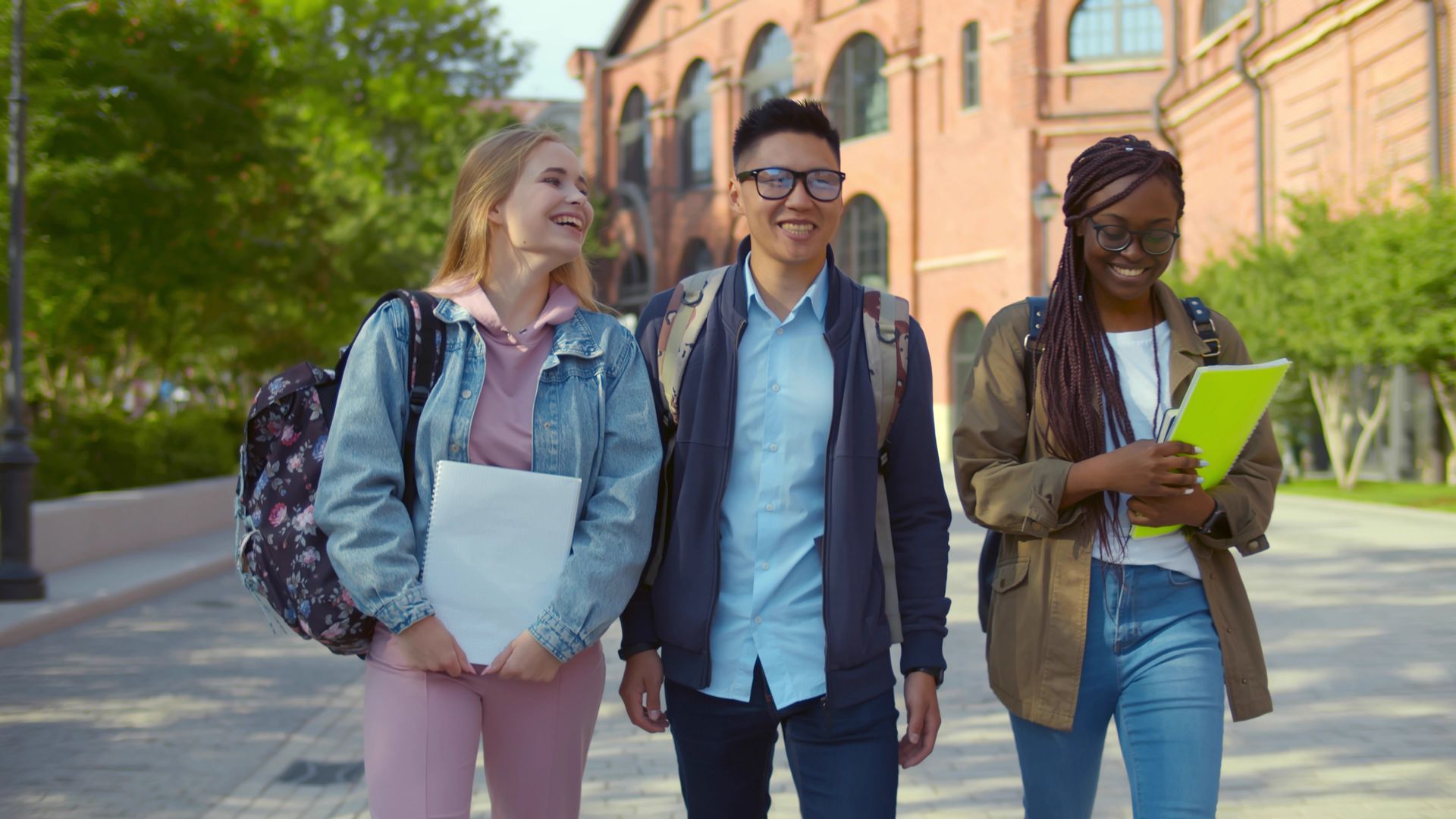 Three students walking and smiling outside a brick building, holding books and backpacks.