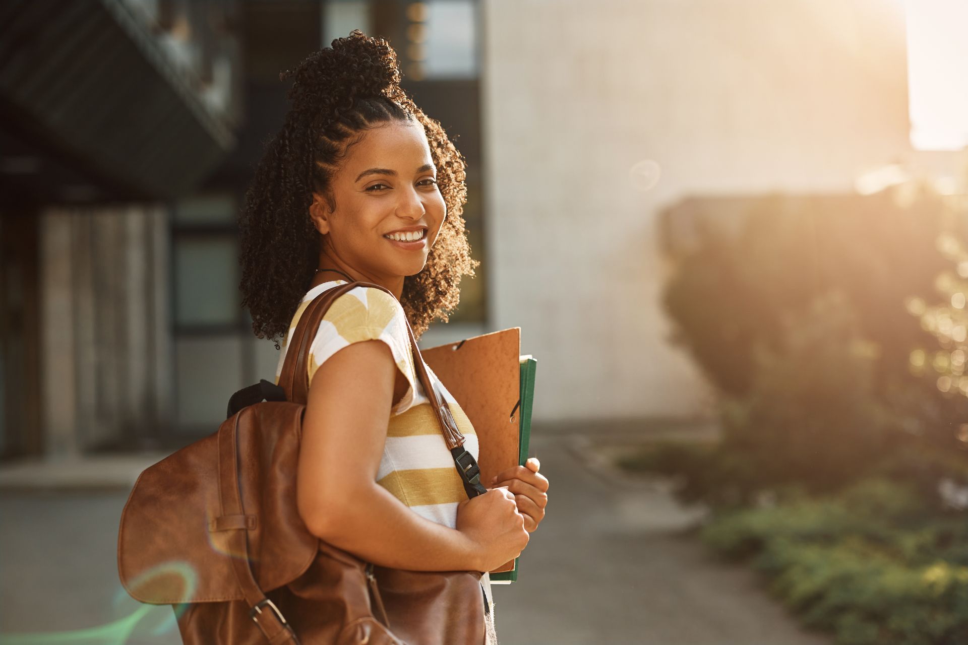 Woman with backpack and books smiles in front of a building in sunlight.