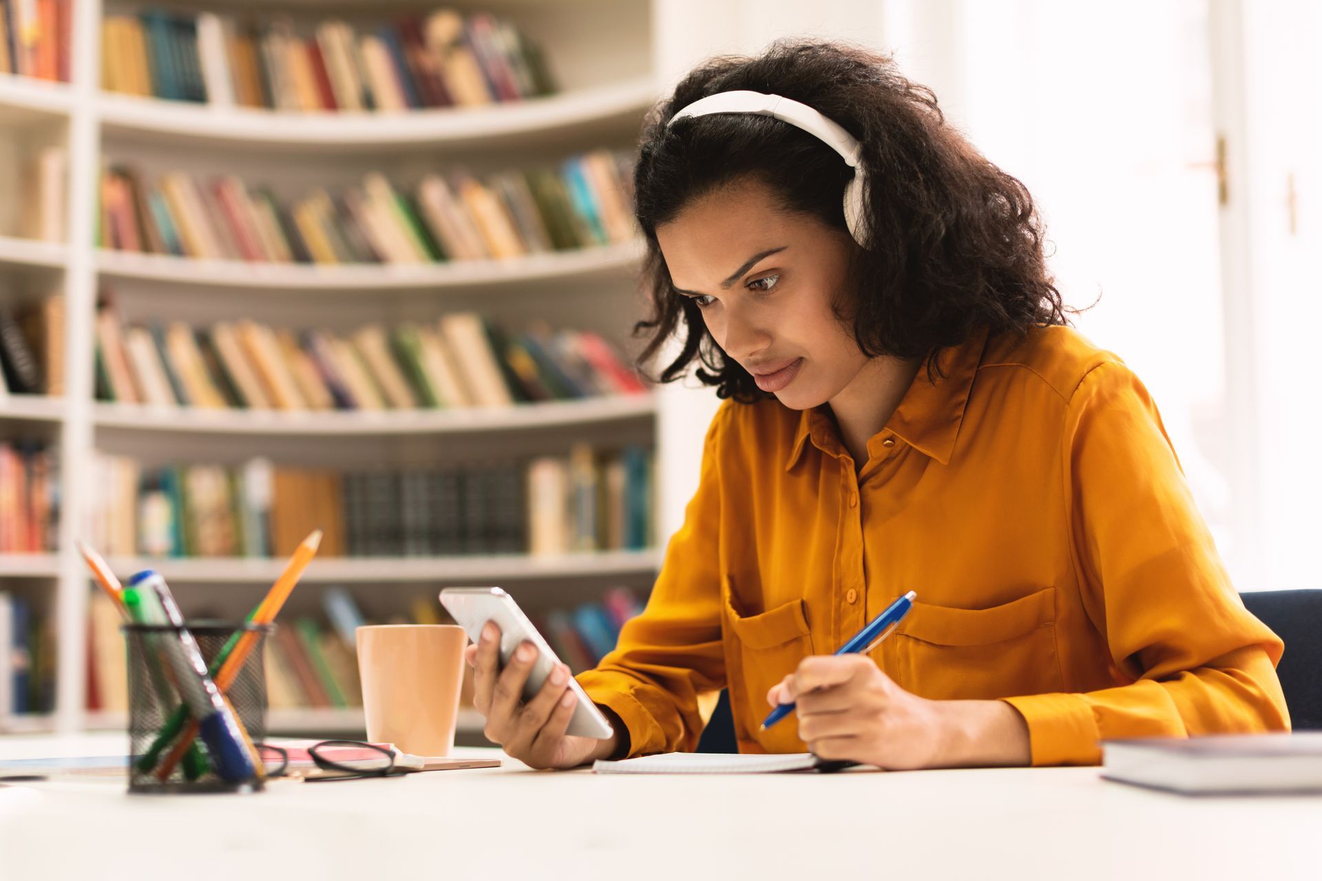 Woman wearing headphones, using a phone and taking notes at a desk in front of a bookshelf.