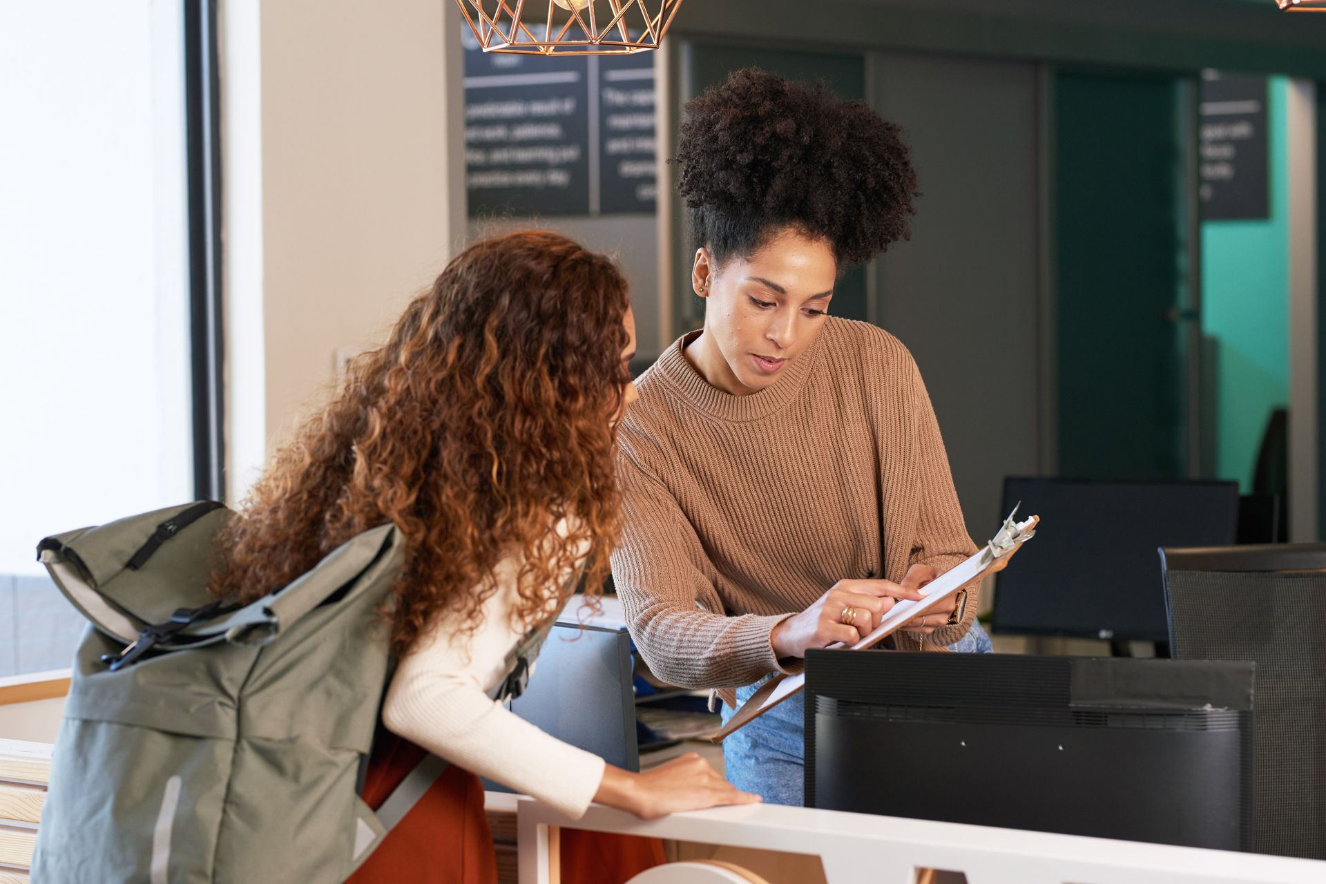 Two women at a counter looking at a clipboard. One is holding the clipboard, the other has a backpack. Two women at a counter looking at a clipboard. One is holding the clipboard, the other has a backpack.