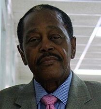 Man in a gray suit, pink tie, and blue collared shirt; he is looking forward.